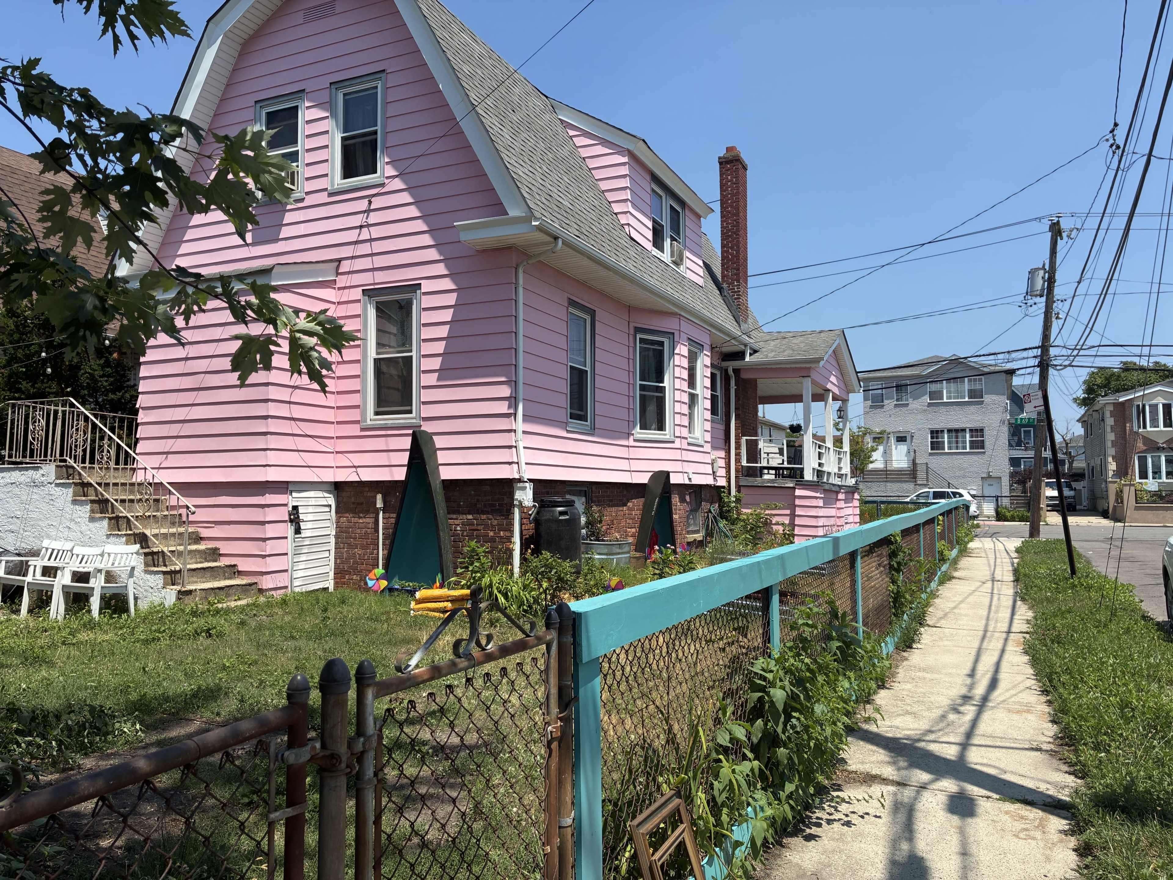 A pink two-story house with a front porch sits next to a sidewalk, surrounded by a yard and a chain-link fence.