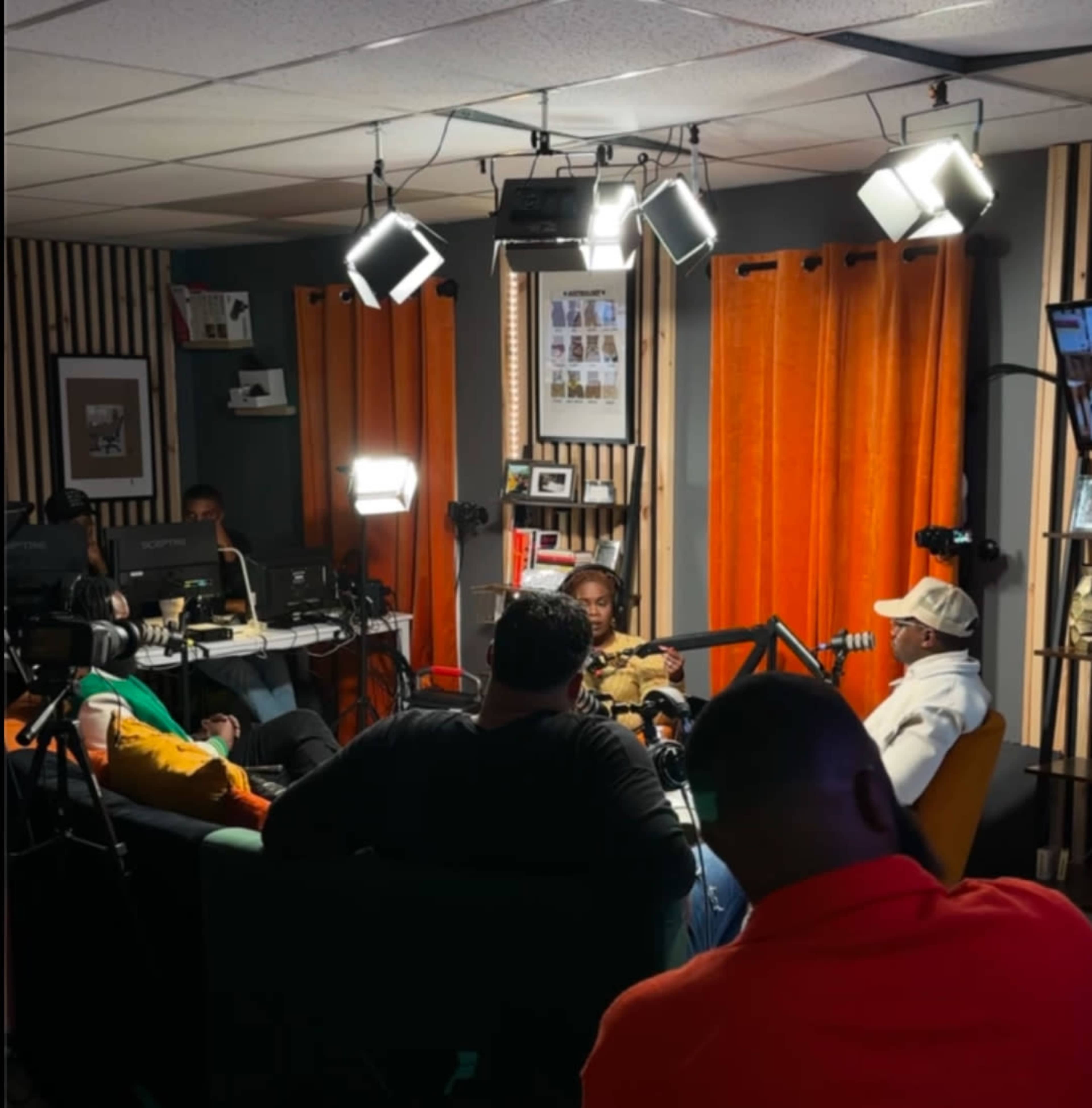 A group of people sits in a studio with bright orange curtains, engaged in a conversation while being filmed under multiple lights.