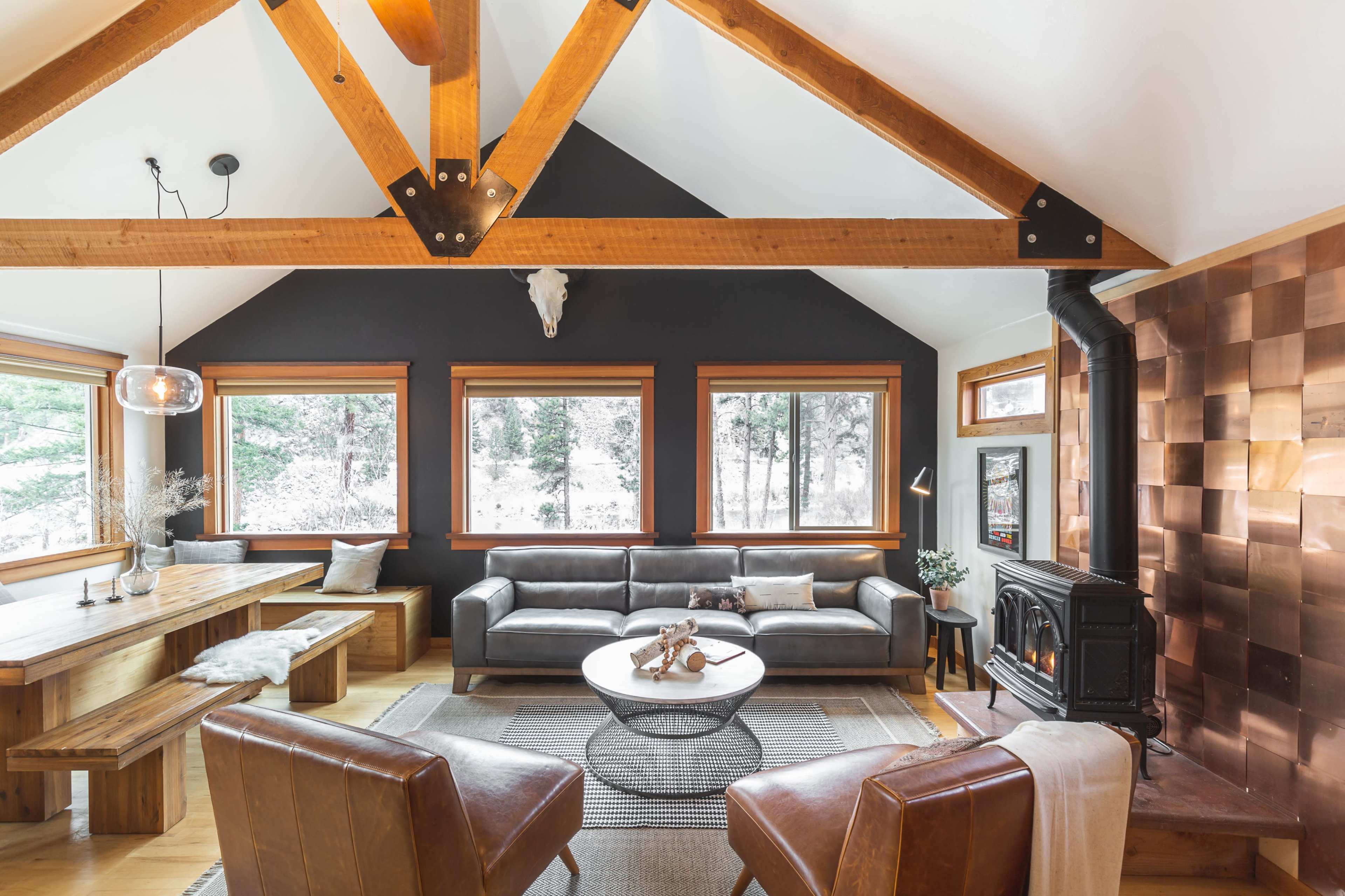 A spacious living room features a wood-beamed ceiling, a leather sofa, a circular coffee table, and large windows overlooking a snowy landscape.
