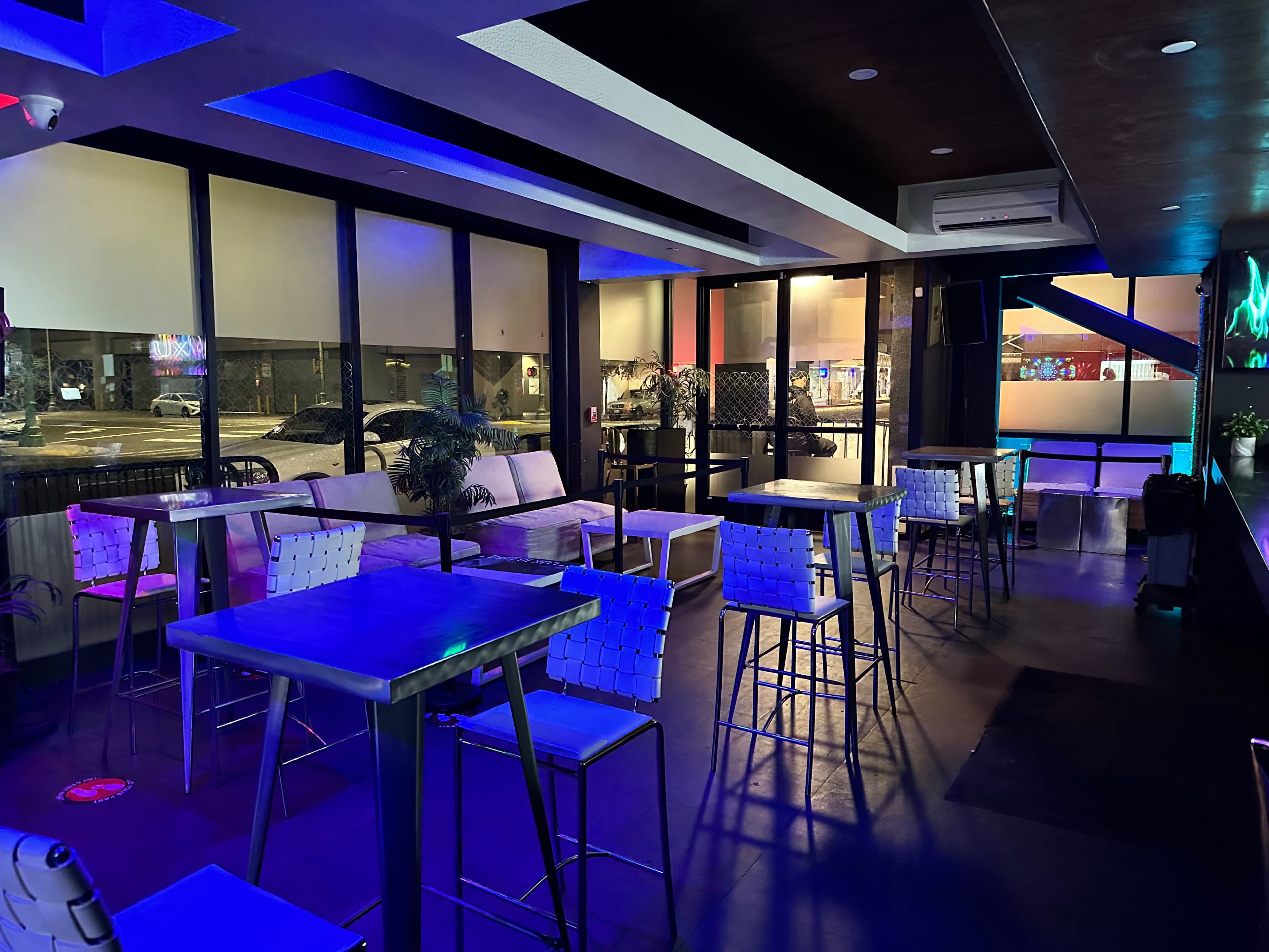 The image shows a dimly lit lounge area with several tall tables and chairs arranged around, and large windows revealing a street view outside.