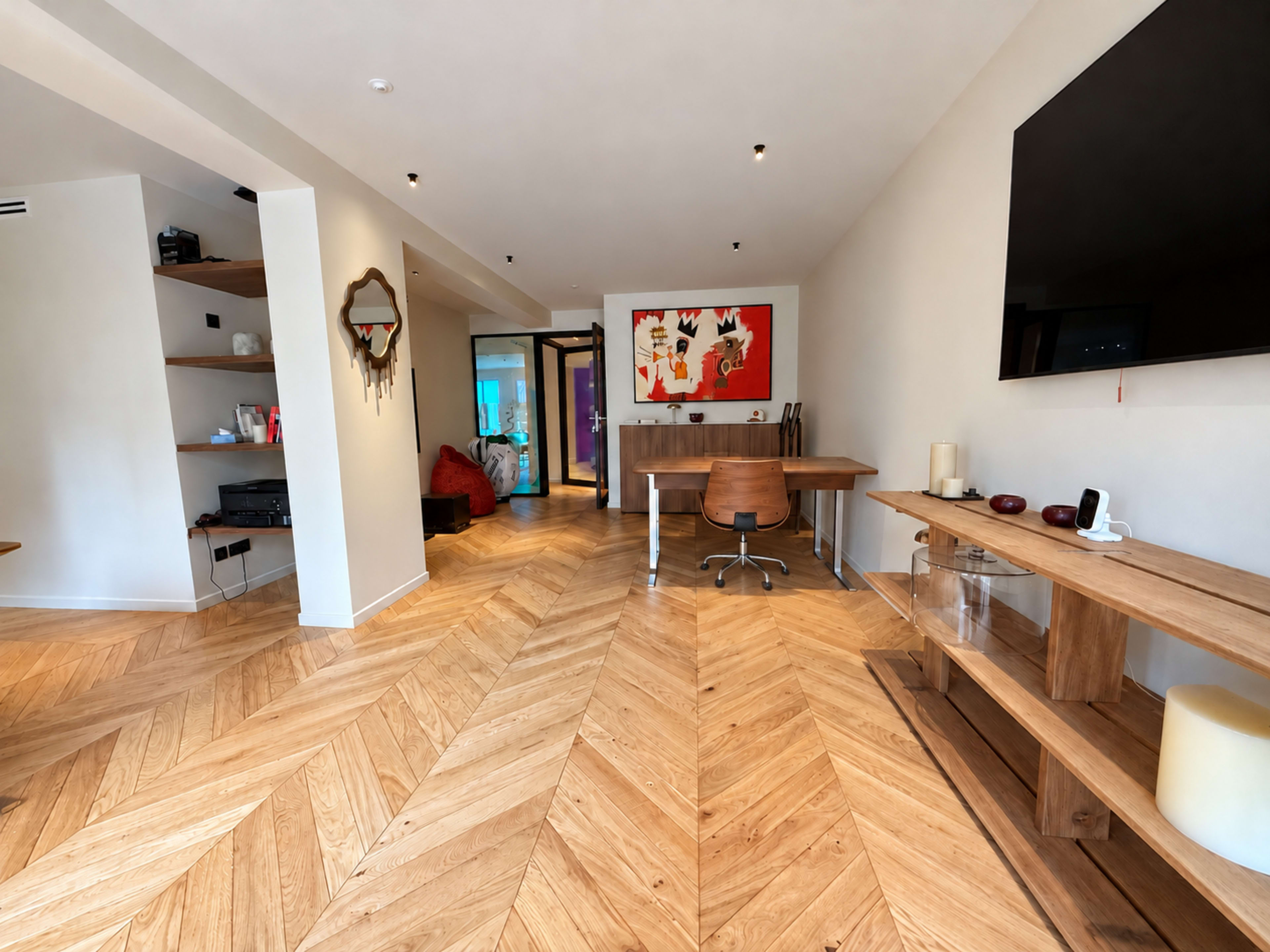 The image shows a modern interior space with wooden chevron flooring, a desk with a chair, and colorful artwork on the wall, leading to other rooms.