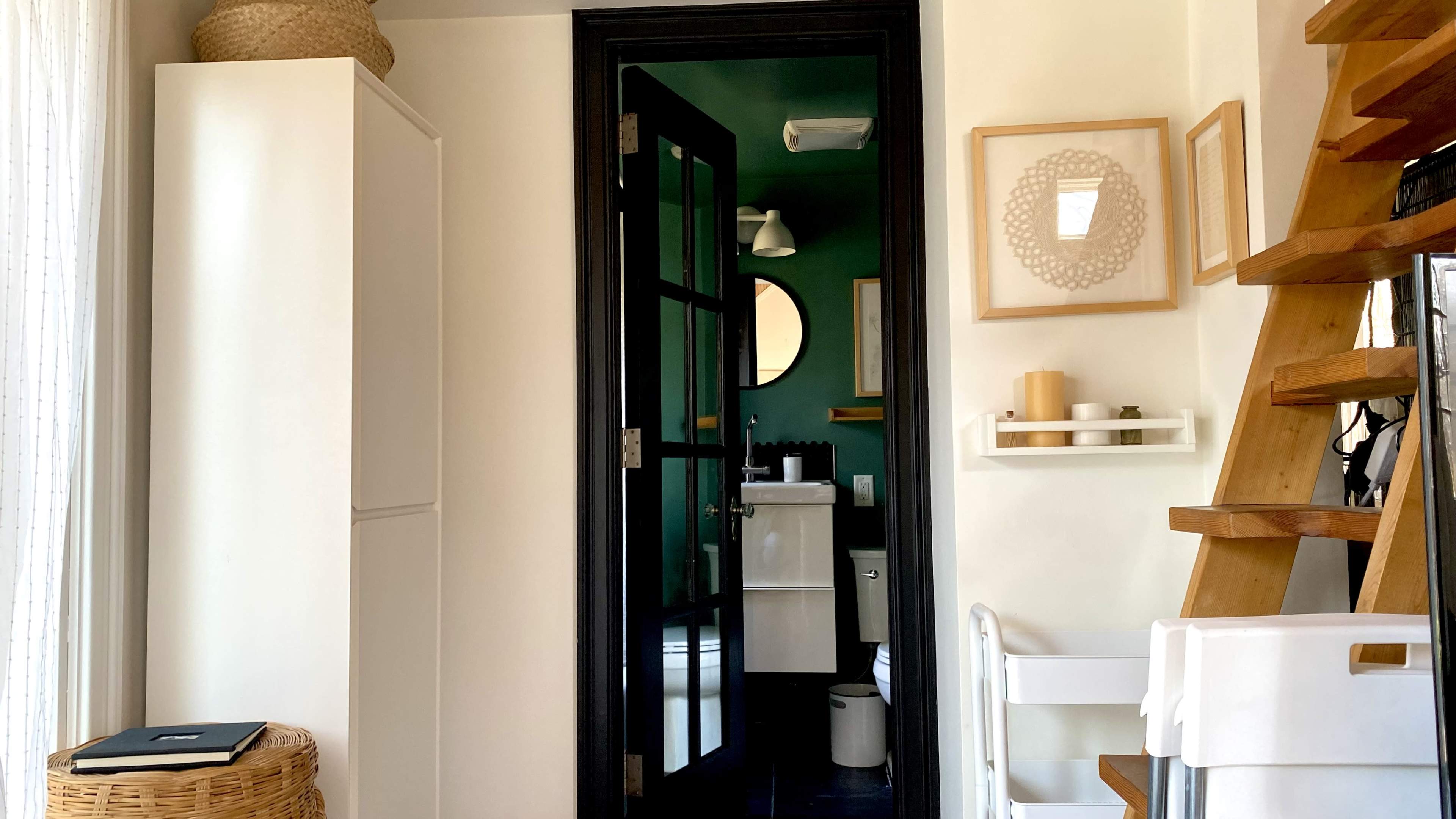 A doorway leads into a bathroom, featuring a black door, green walls, and a circular mirror, with wooden stairs visible on the right.