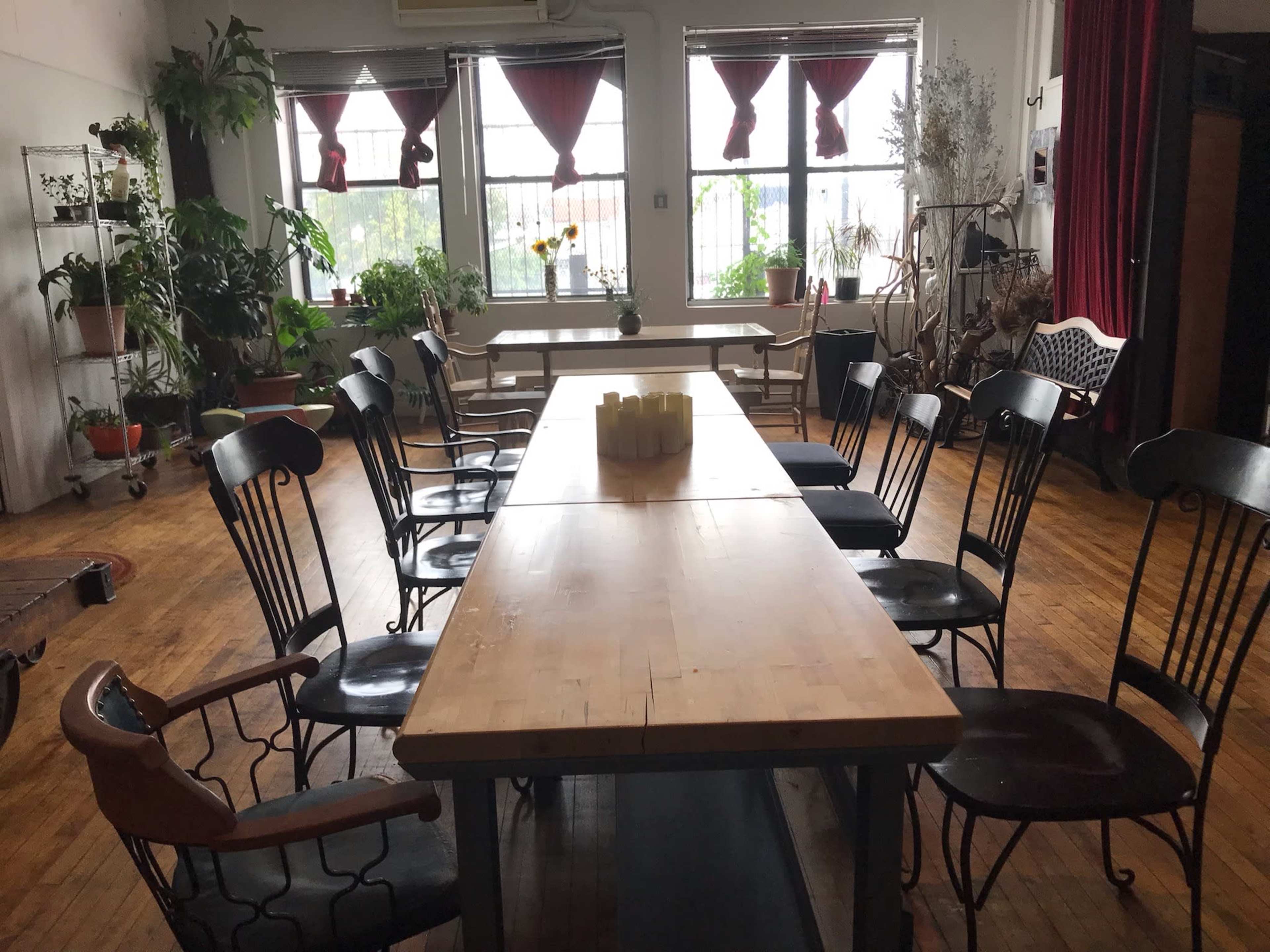 A long wooden table surrounded by various chairs is set up in a room with large windows and several plants.