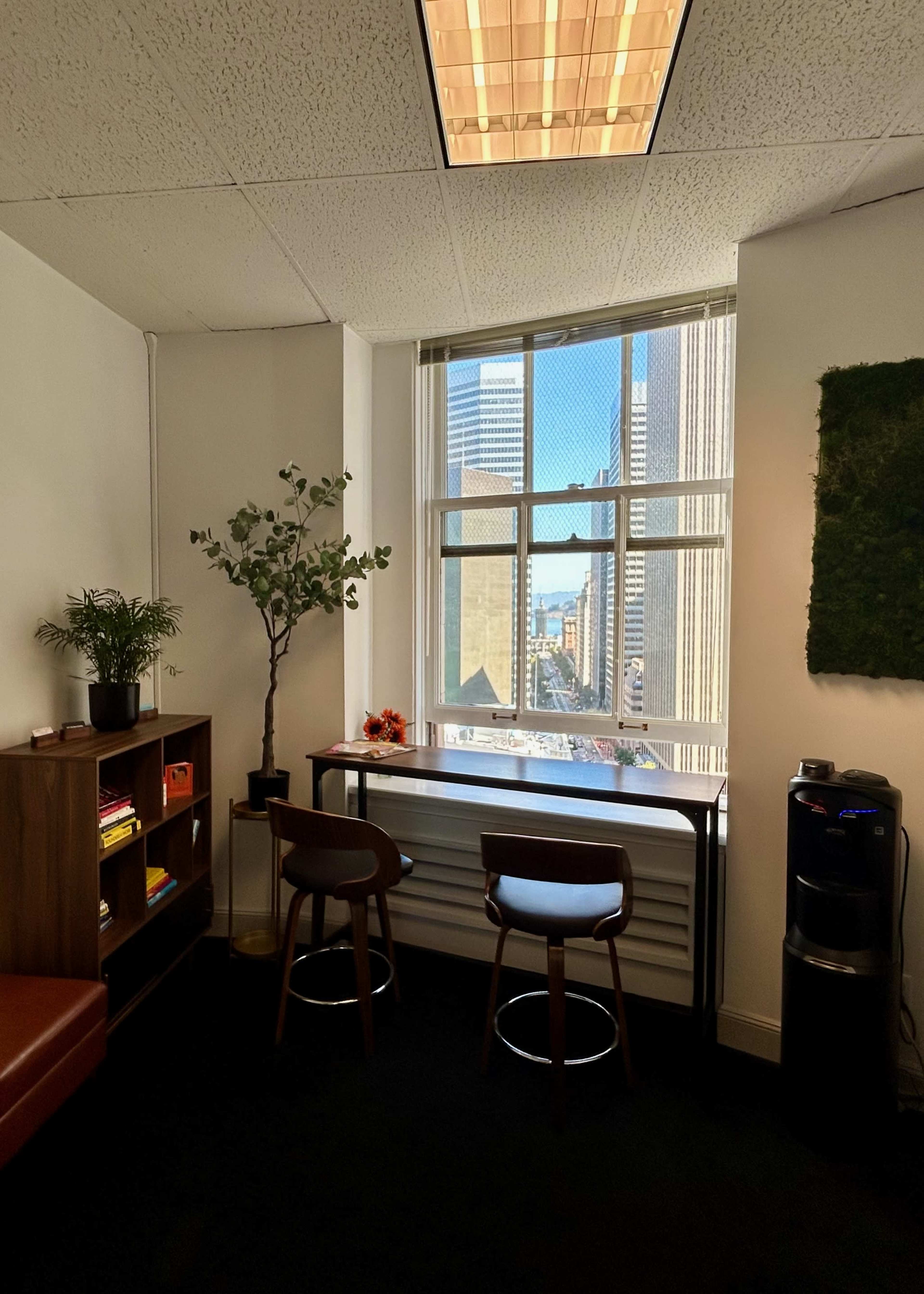 An office corner features a window with a view of a city skyline, two stools by a countertop, and a bookshelf with plants and decor.