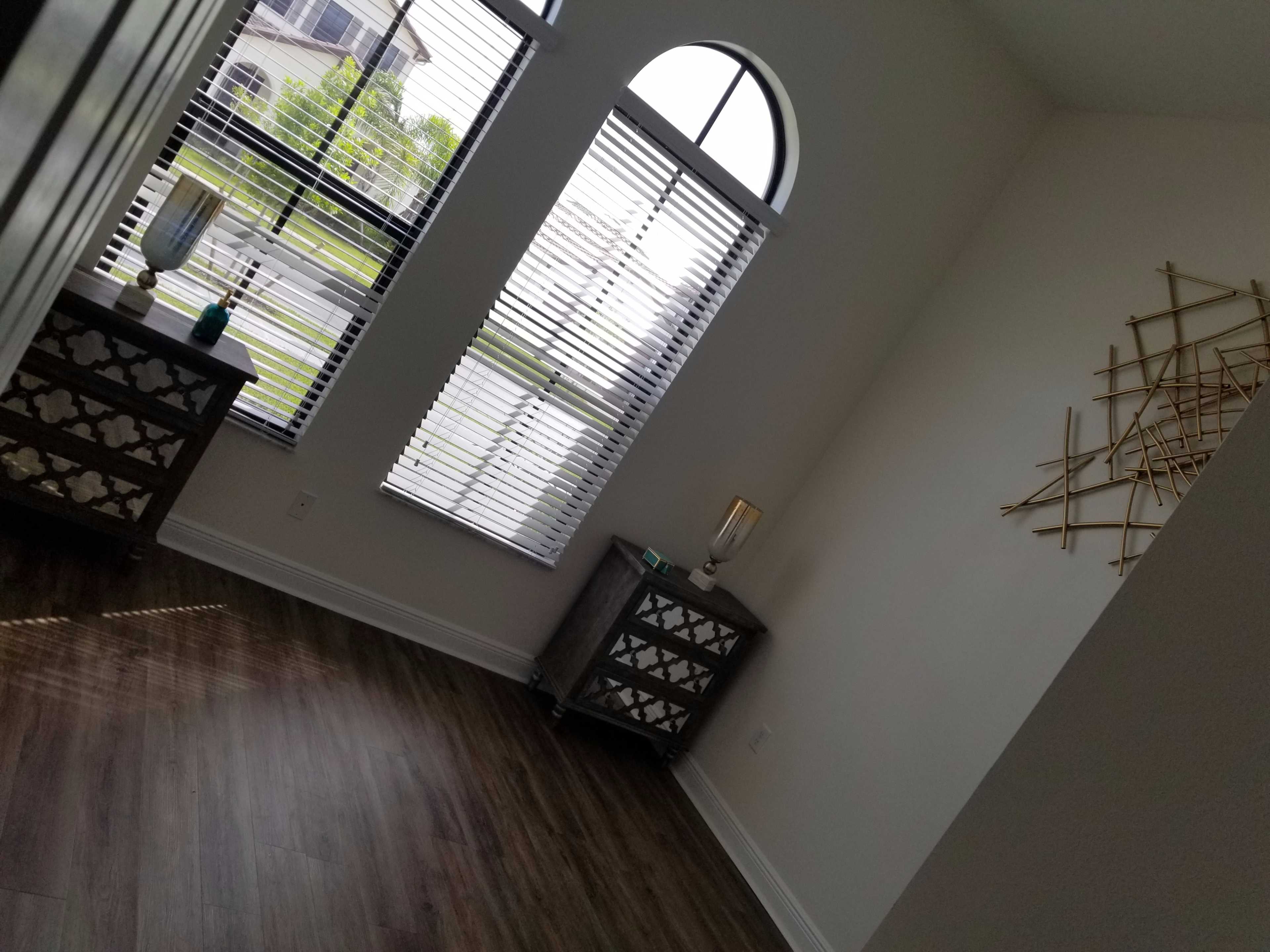 The image shows a corner of a well-lit room featuring two decorative side tables with geometric designs, large windows with blinds, and a wall decor piece.