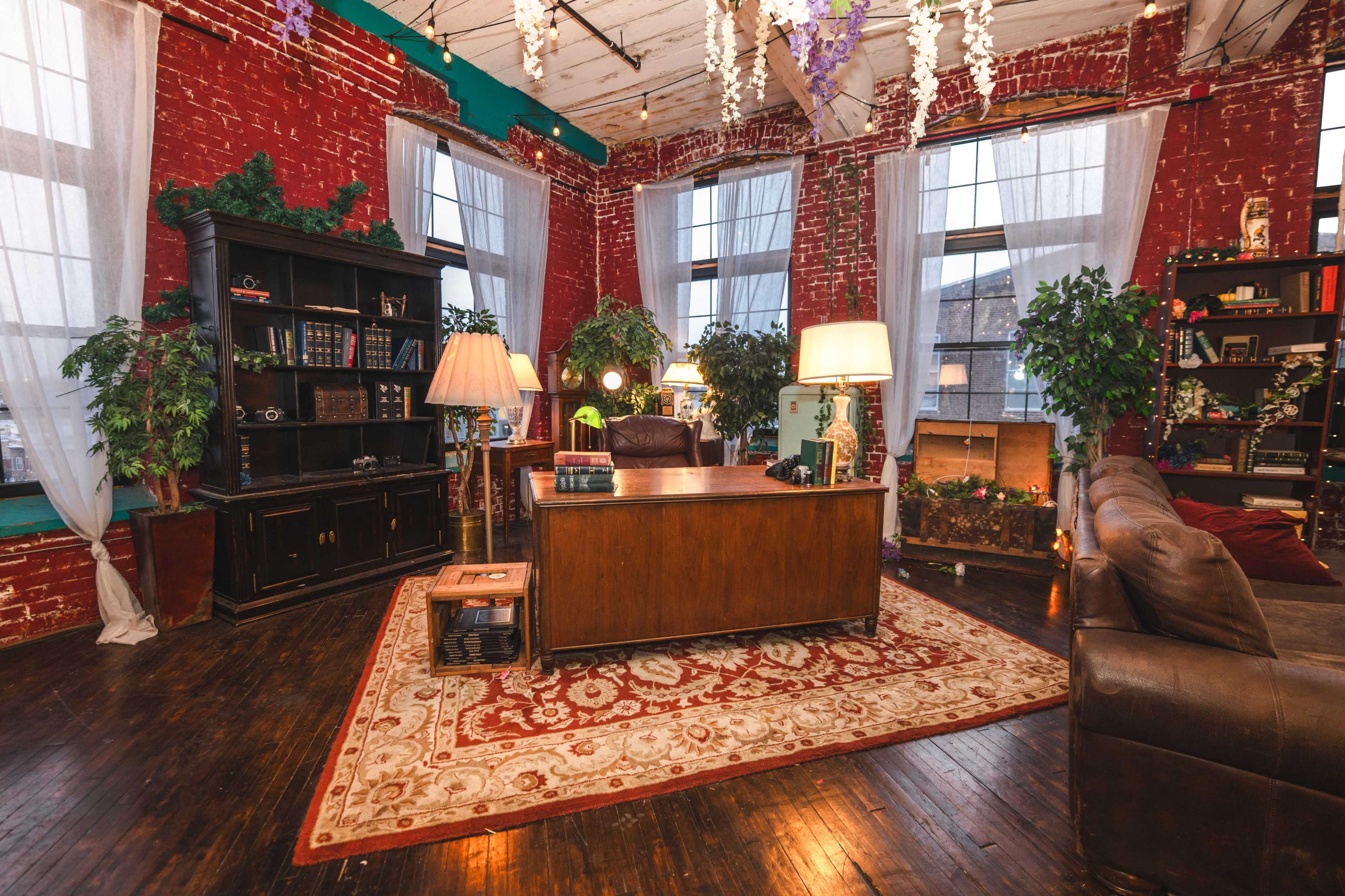 The image shows a cozy office space with red brick walls, a wooden desk, a large bookshelf, plants, and decorative lighting.