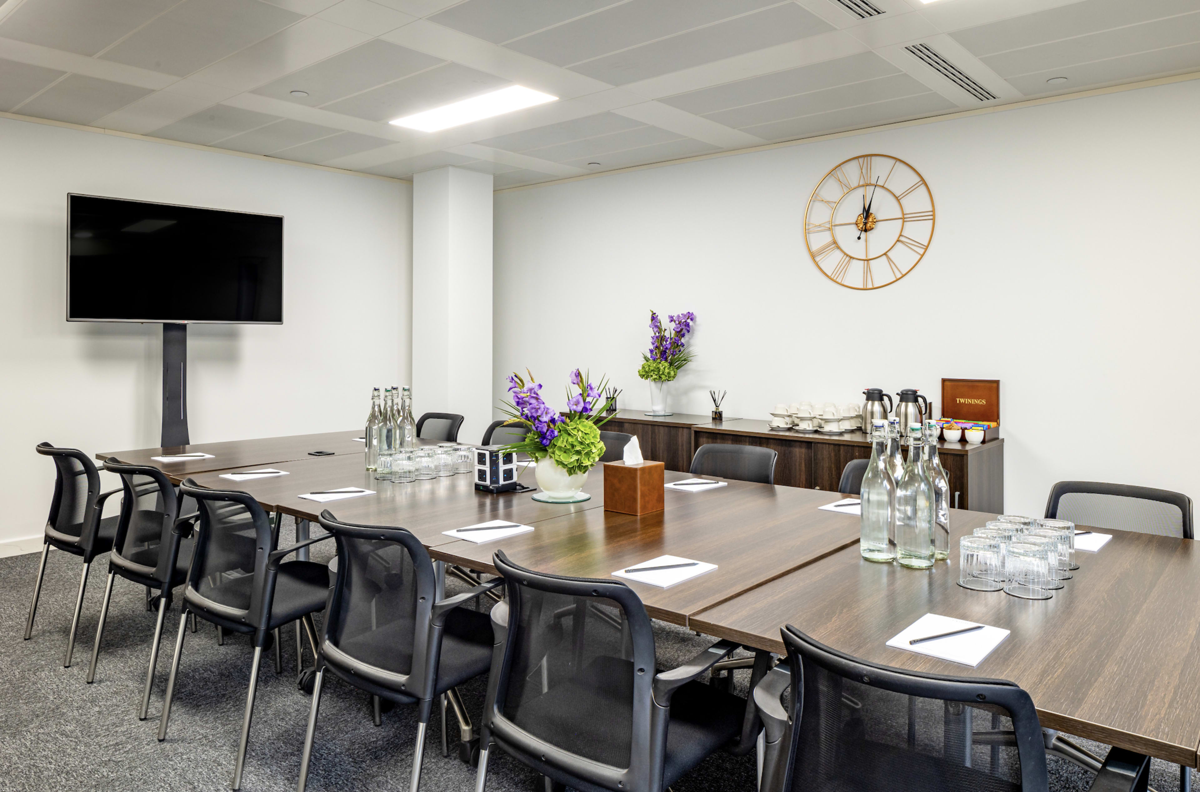 A modern conference room features a long table surrounded by chairs, with a television mounted on the wall and a clock above a sideboard.