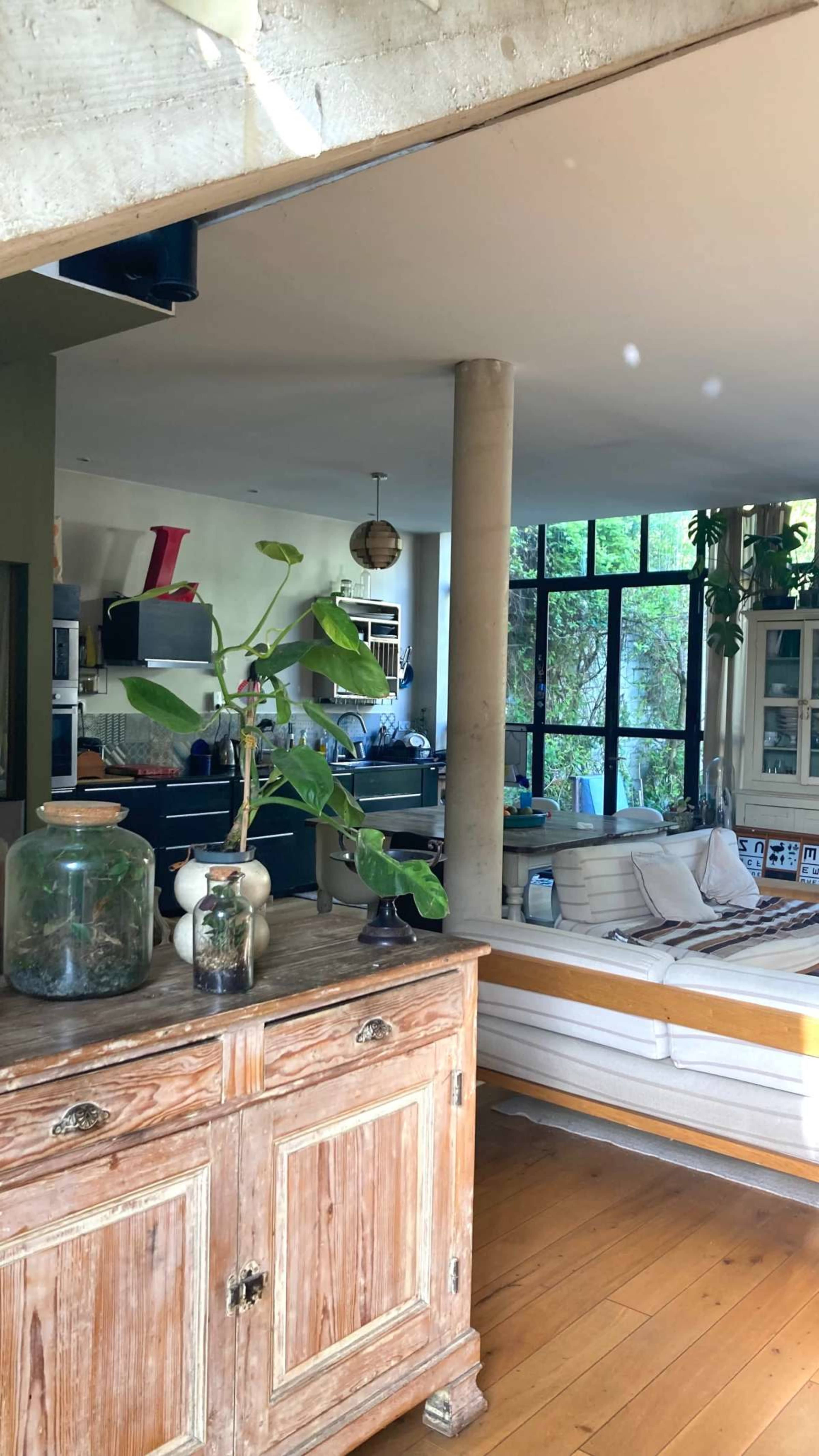 The image shows an interior space with wooden flooring, a vintage cabinet, and a seating area with a sofa, surrounded by large windows and greenery.