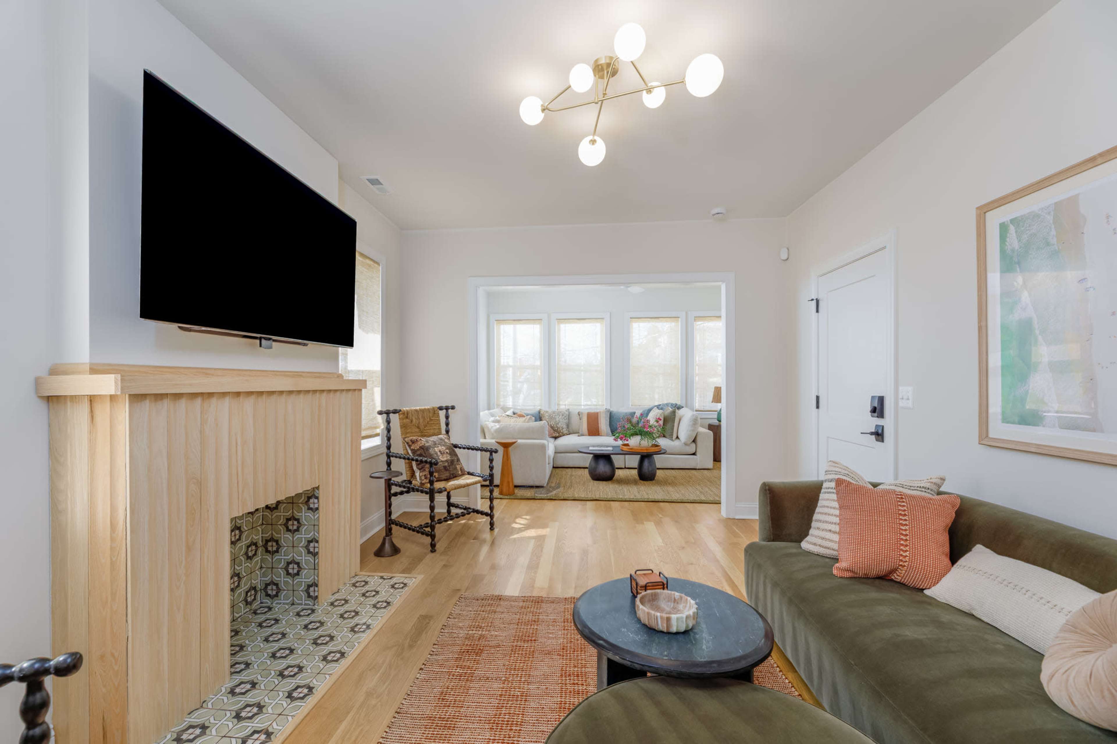The image shows a modern living room featuring a green sofa, a round black coffee table, and a mounted television, with an adjacent sunlit room visible through a doorway.