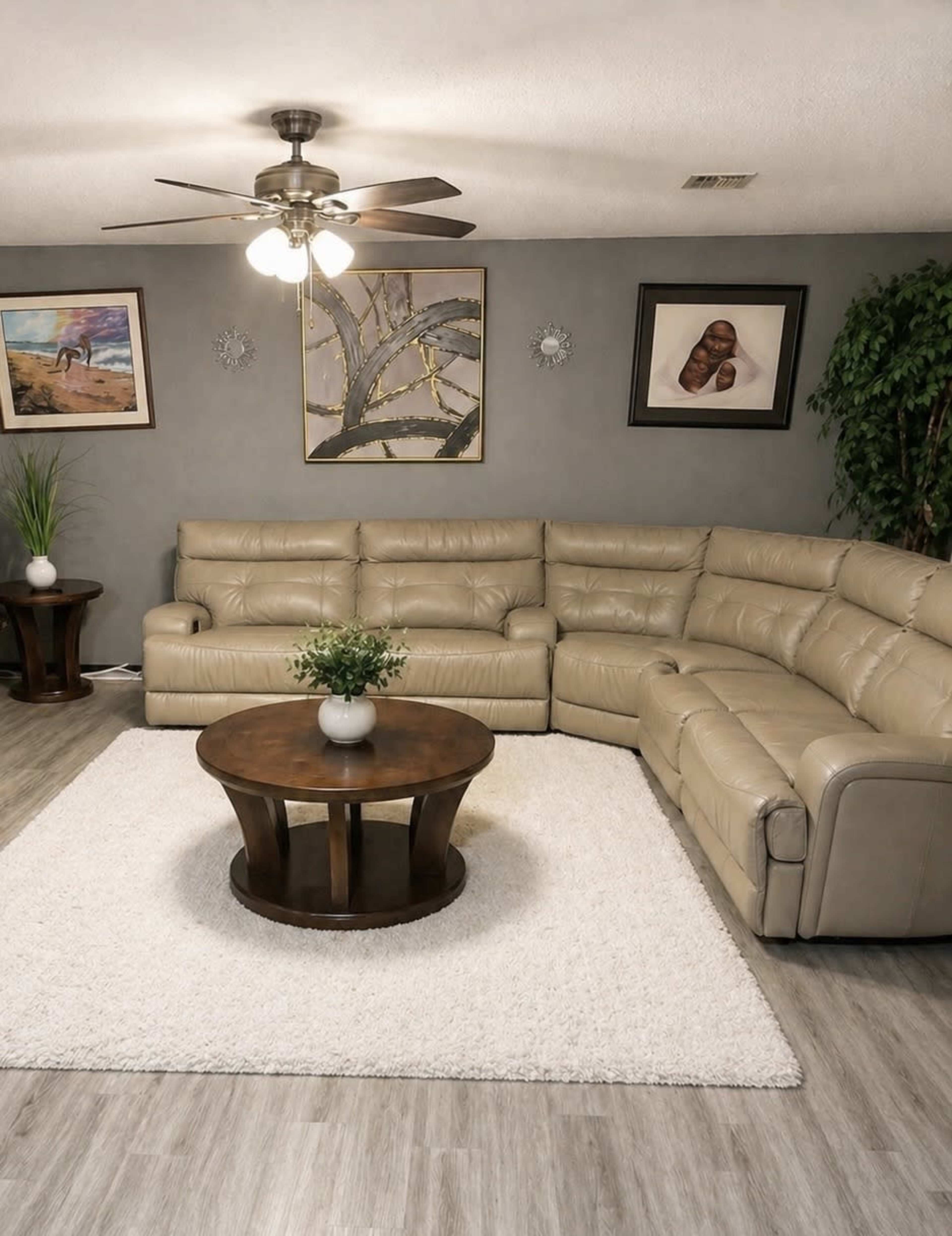 The image shows a modern living room featuring a light beige sectional sofa, a round wooden coffee table, and a white area rug, with art and plants displayed on the walls.