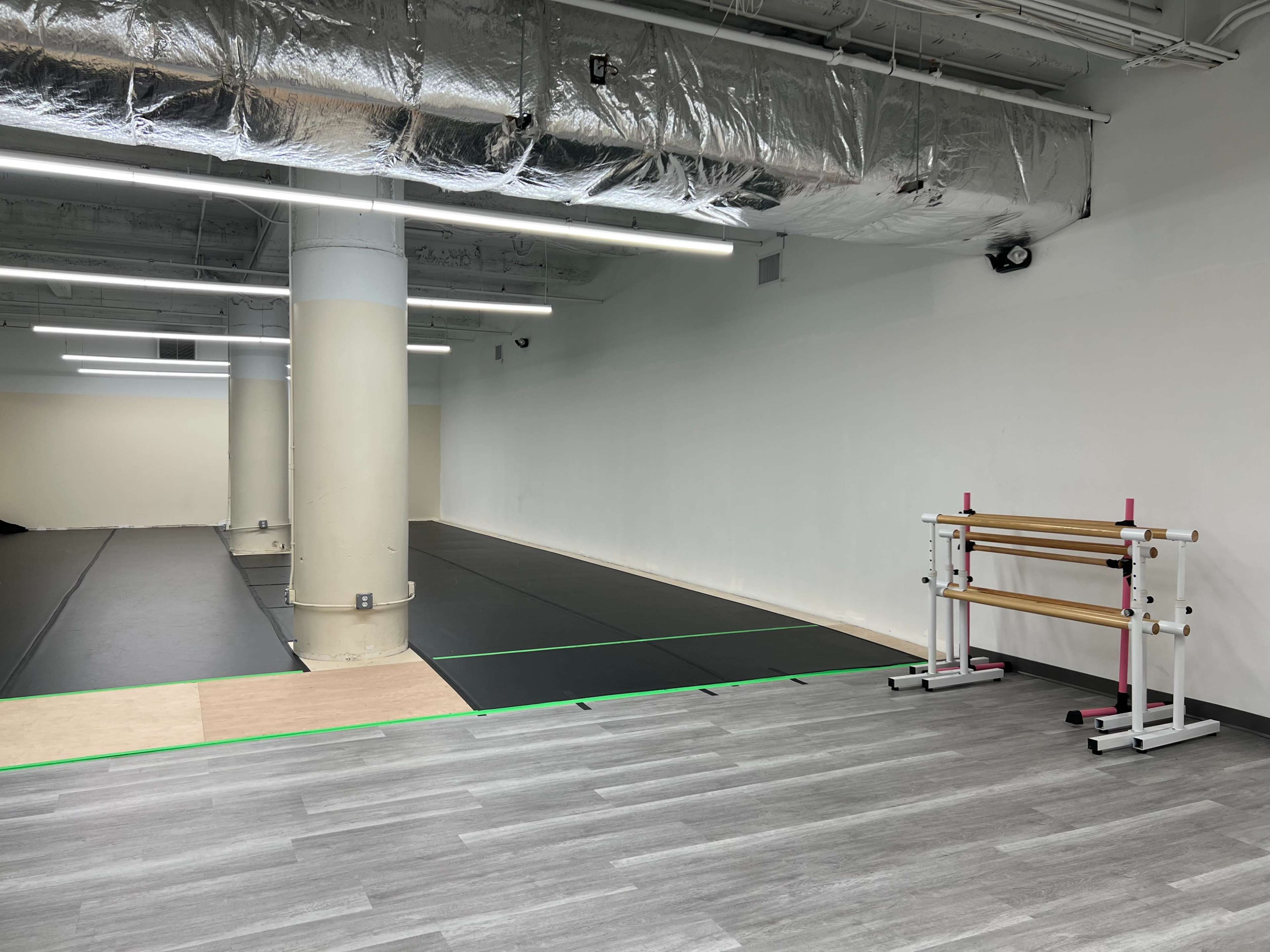 A spacious, empty gym area featuring black mats on the floor, a white wall, and a wooden ballet barre against the side.