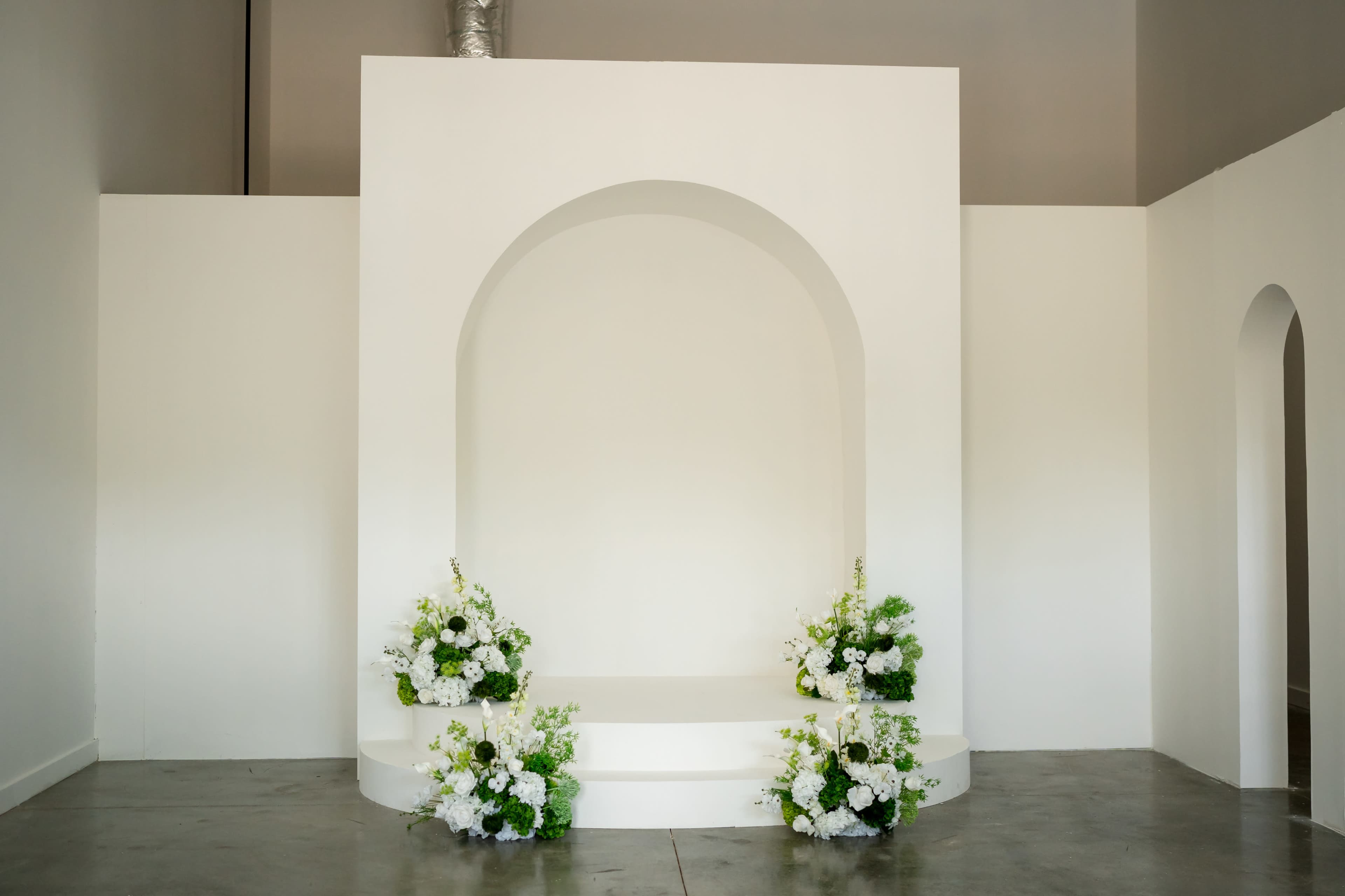The image shows a minimalist white stage with an arched backdrop, flanked by floral arrangements at the base.