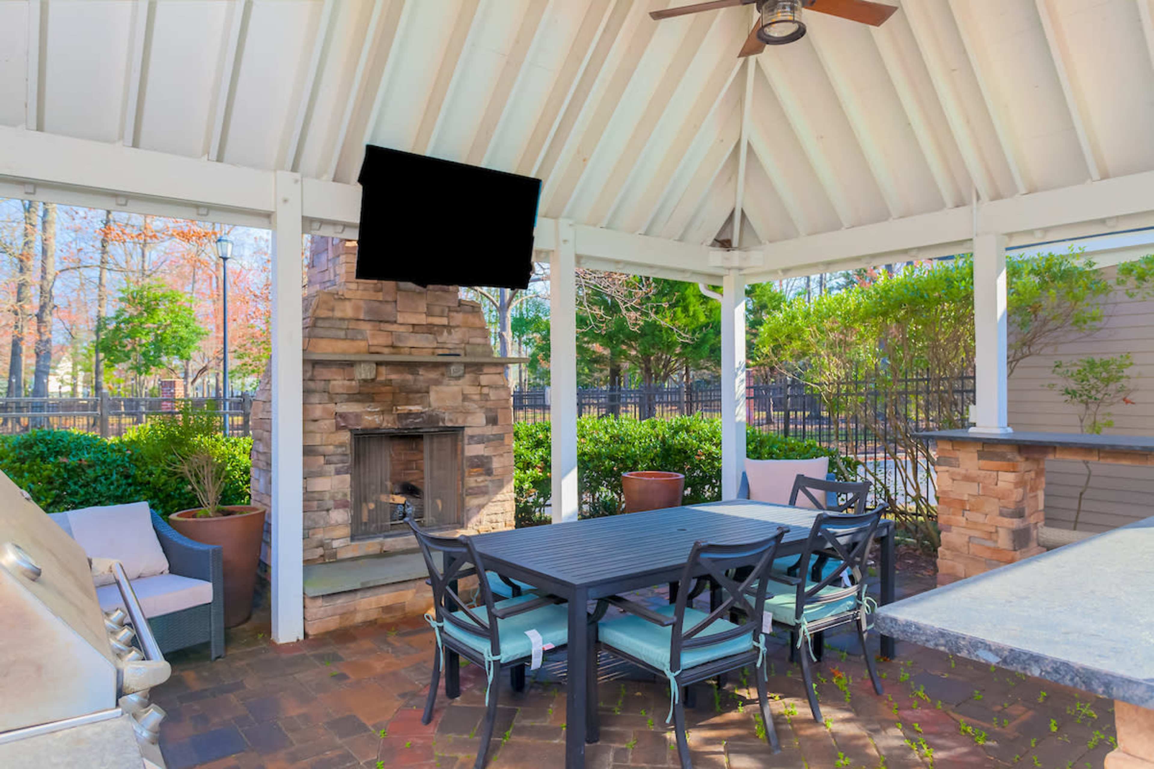 A covered outdoor patio features a stone fireplace and a dining table with chairs, surrounded by greenery and a barbecue grill.