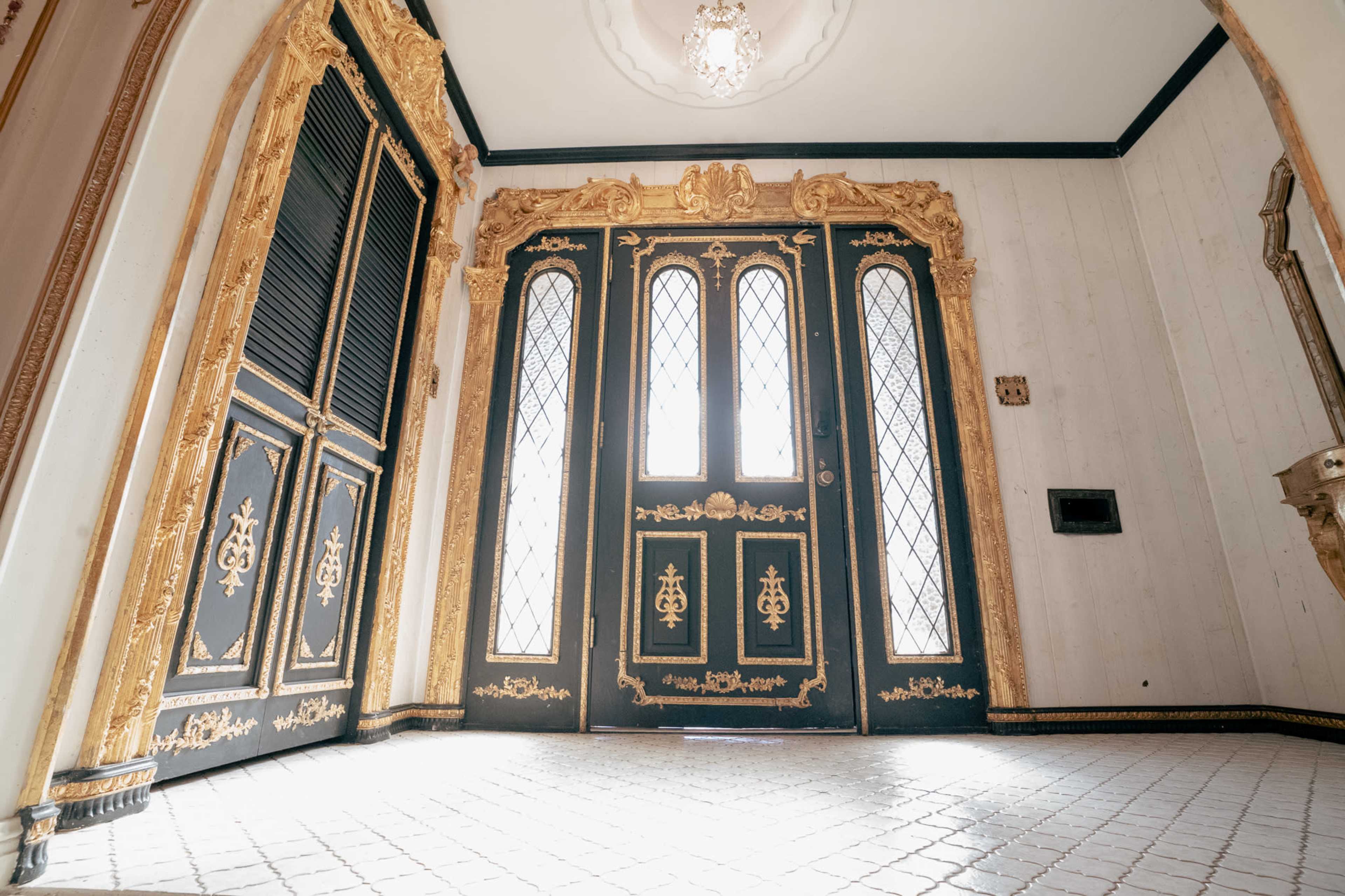 The image shows an ornate entrance with two large doors featuring intricate gold designs and diamond-patterned glass panels, illuminated by a chandelier above.