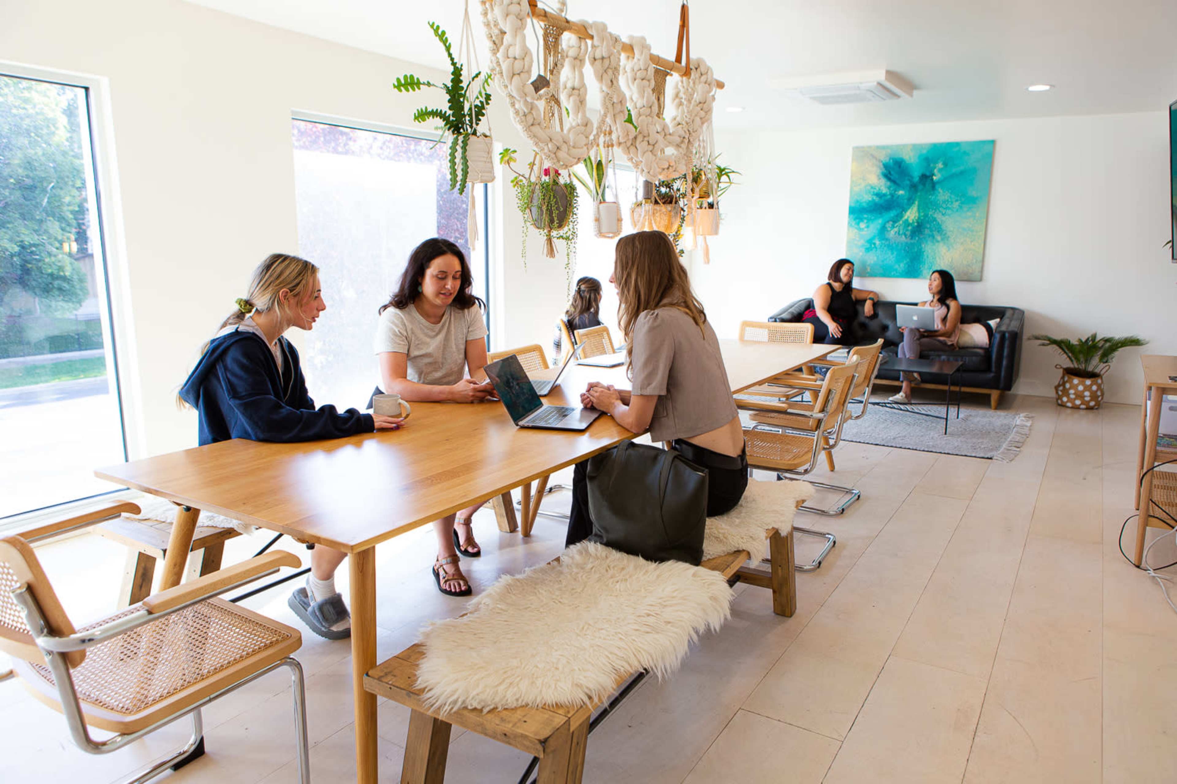 A group of women are gathered in a bright, modern workspace, engaging in conversation and using laptops around a wooden table.