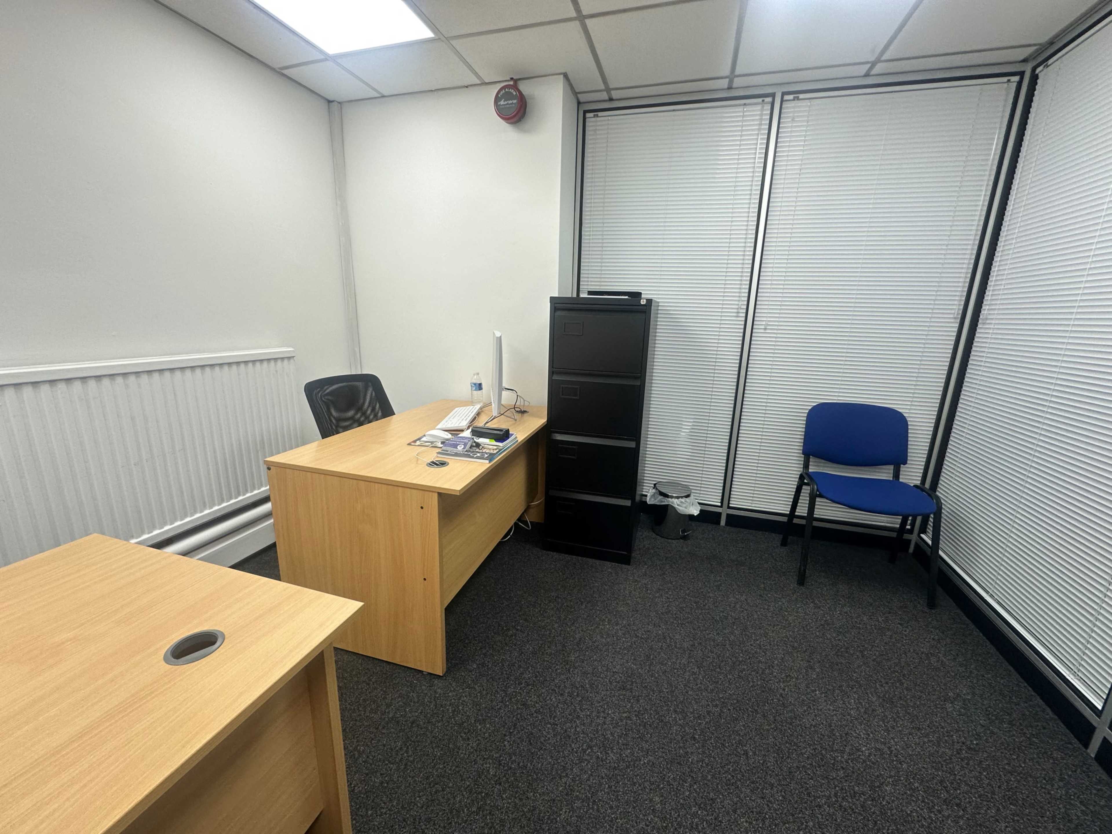 The image shows a small office with a wooden desk, an office chair, a filing cabinet, and a single blue chair, all set against a backdrop of white walls and window blinds.