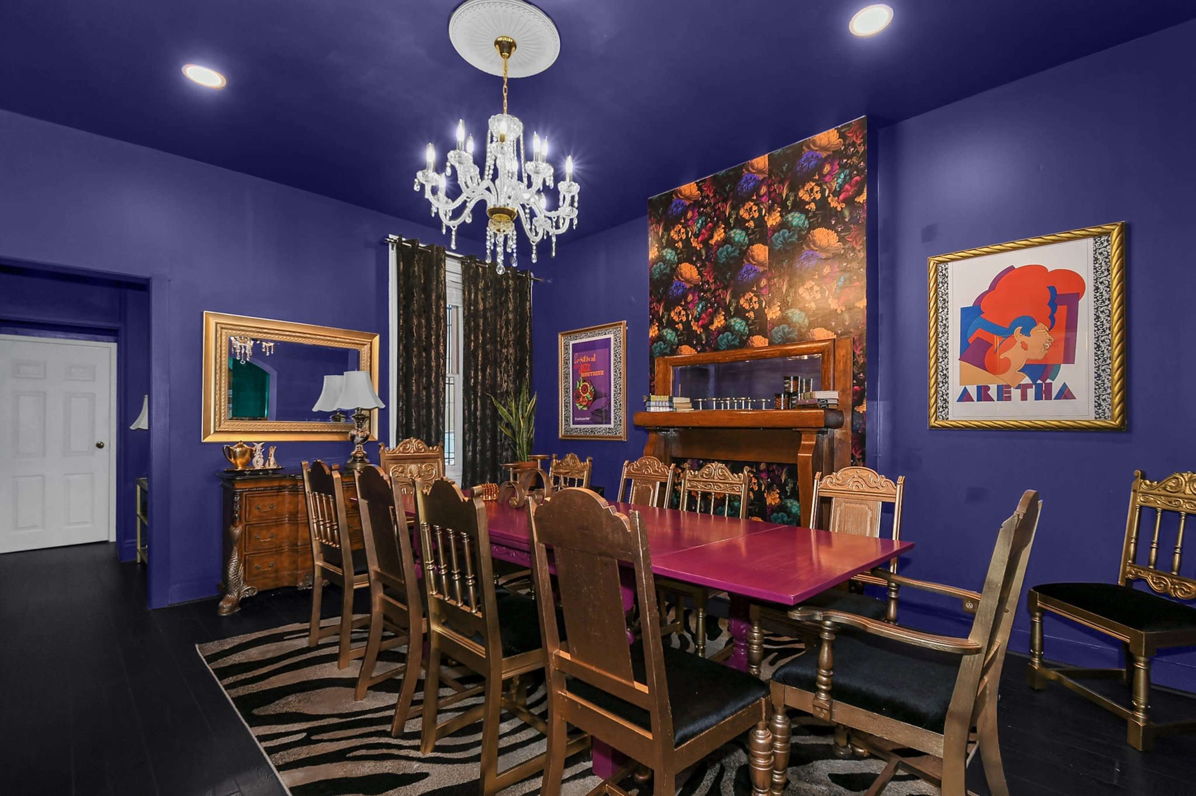A dining room features a purple wall, a large table with ornate chairs, a chandelier, and artistic decor.