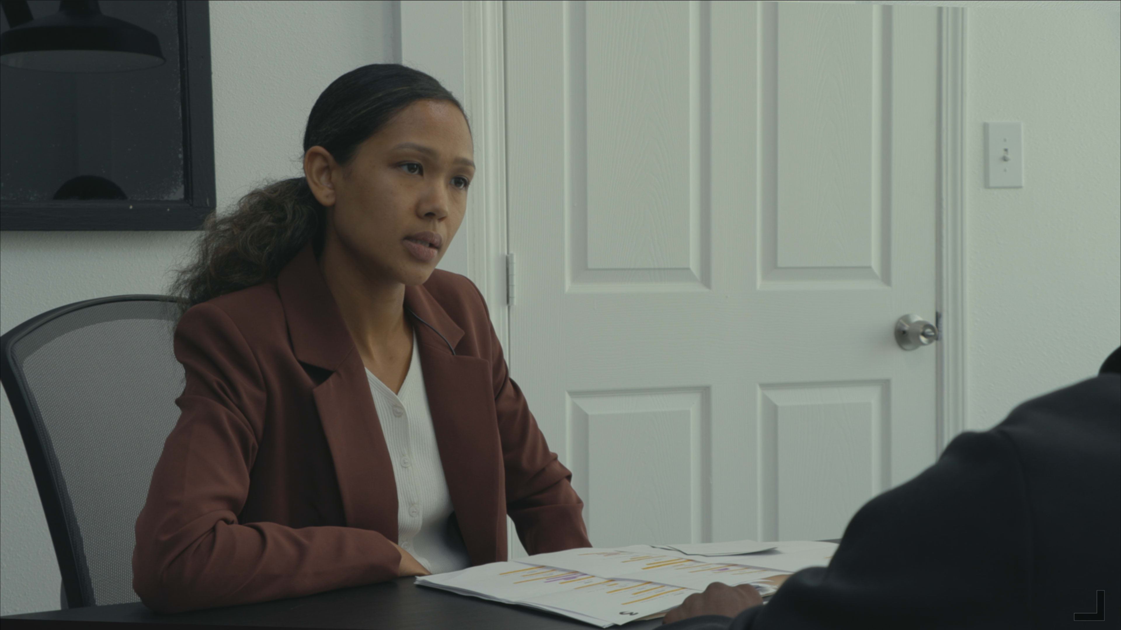 A woman in a brown blazer sits at a table, looking attentively at a person across from her while reviewing documents.