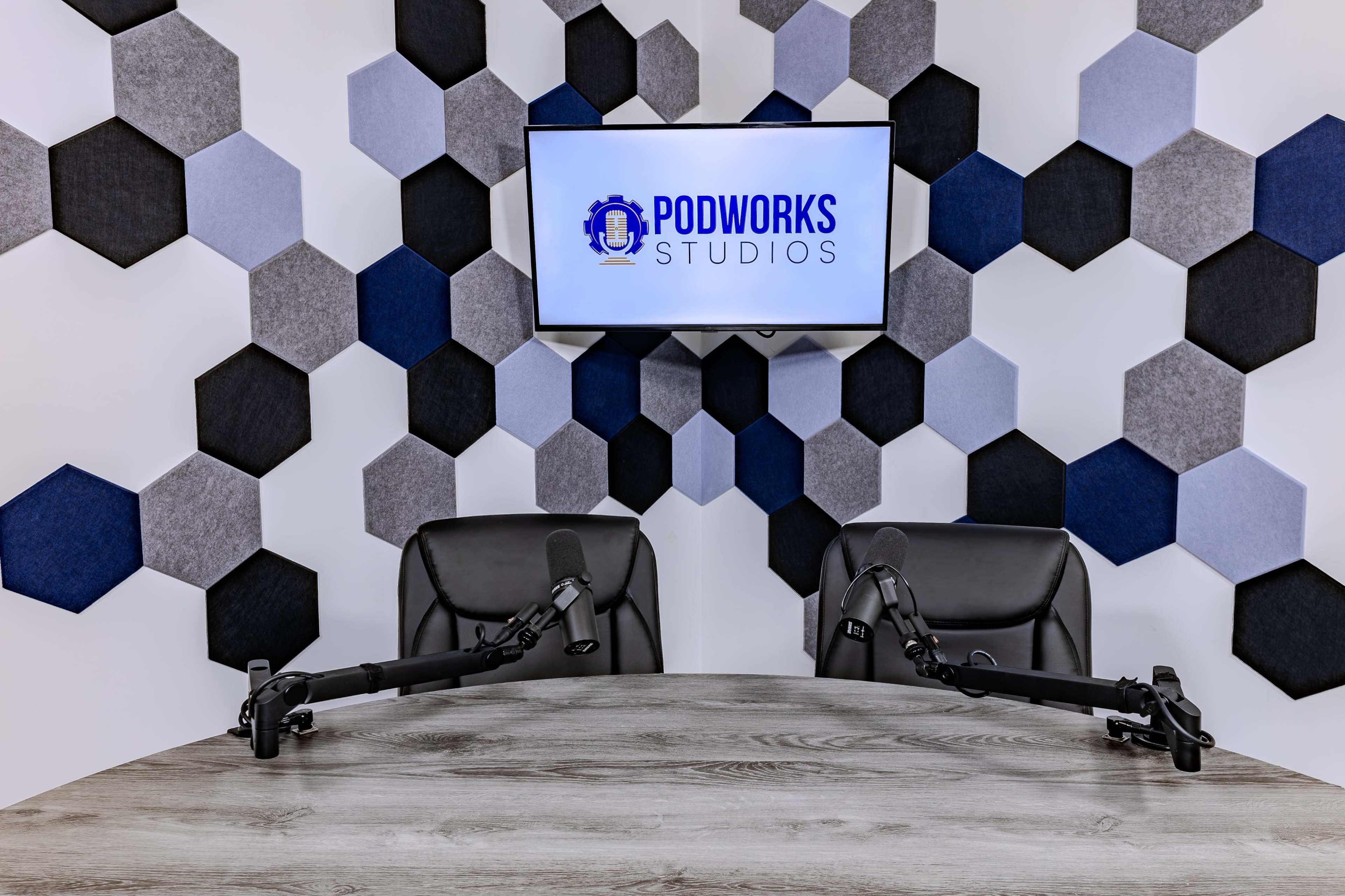 A modern podcast studio features a hexagonal patterned wall, a round table with two chairs, and mounted microphones, with a screen displaying the "Podworks Studios" logo in the background.