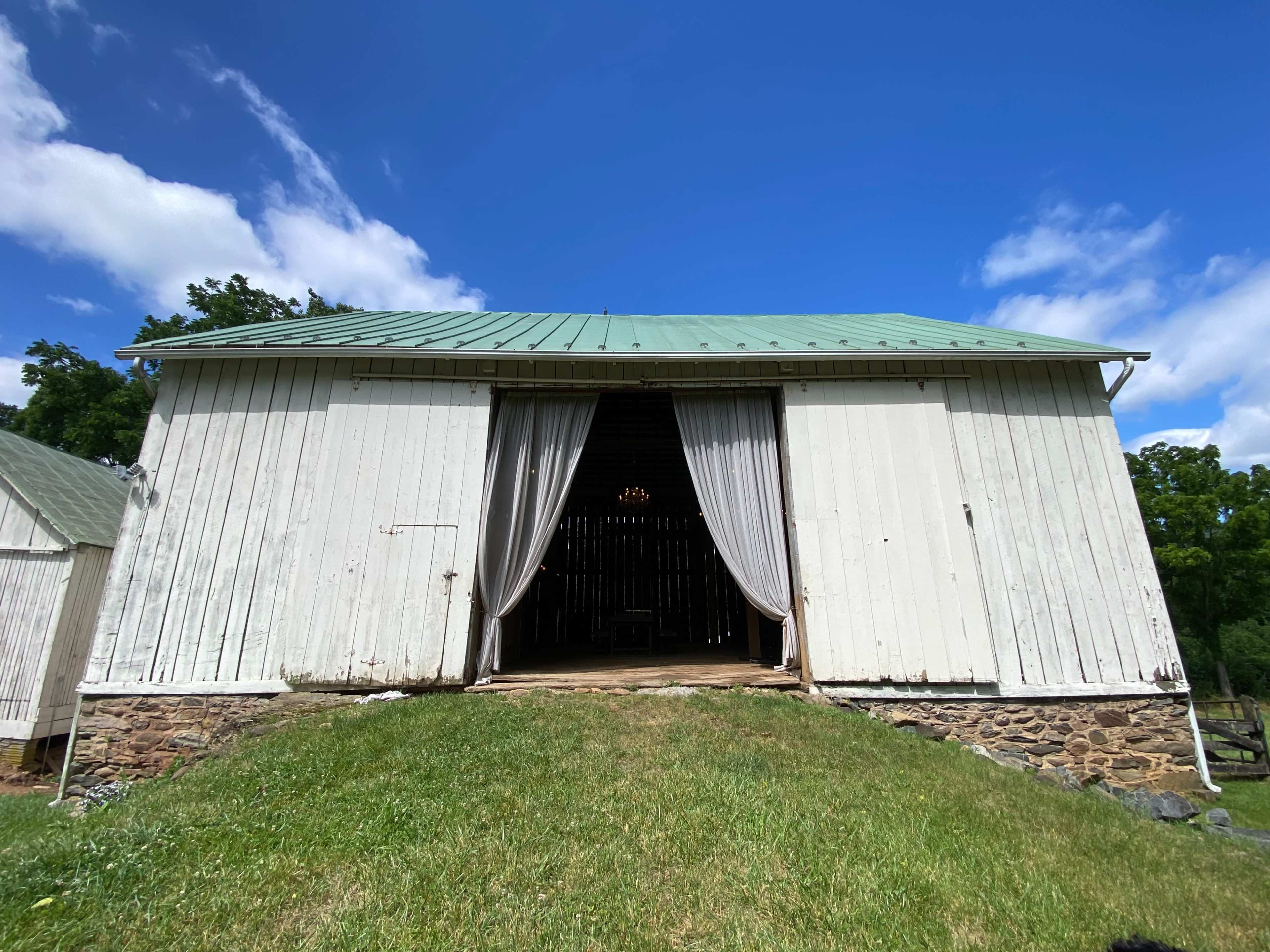 A white barn with a green roof stands on an incline, featuring open curtains at its entrance and a stone foundation.