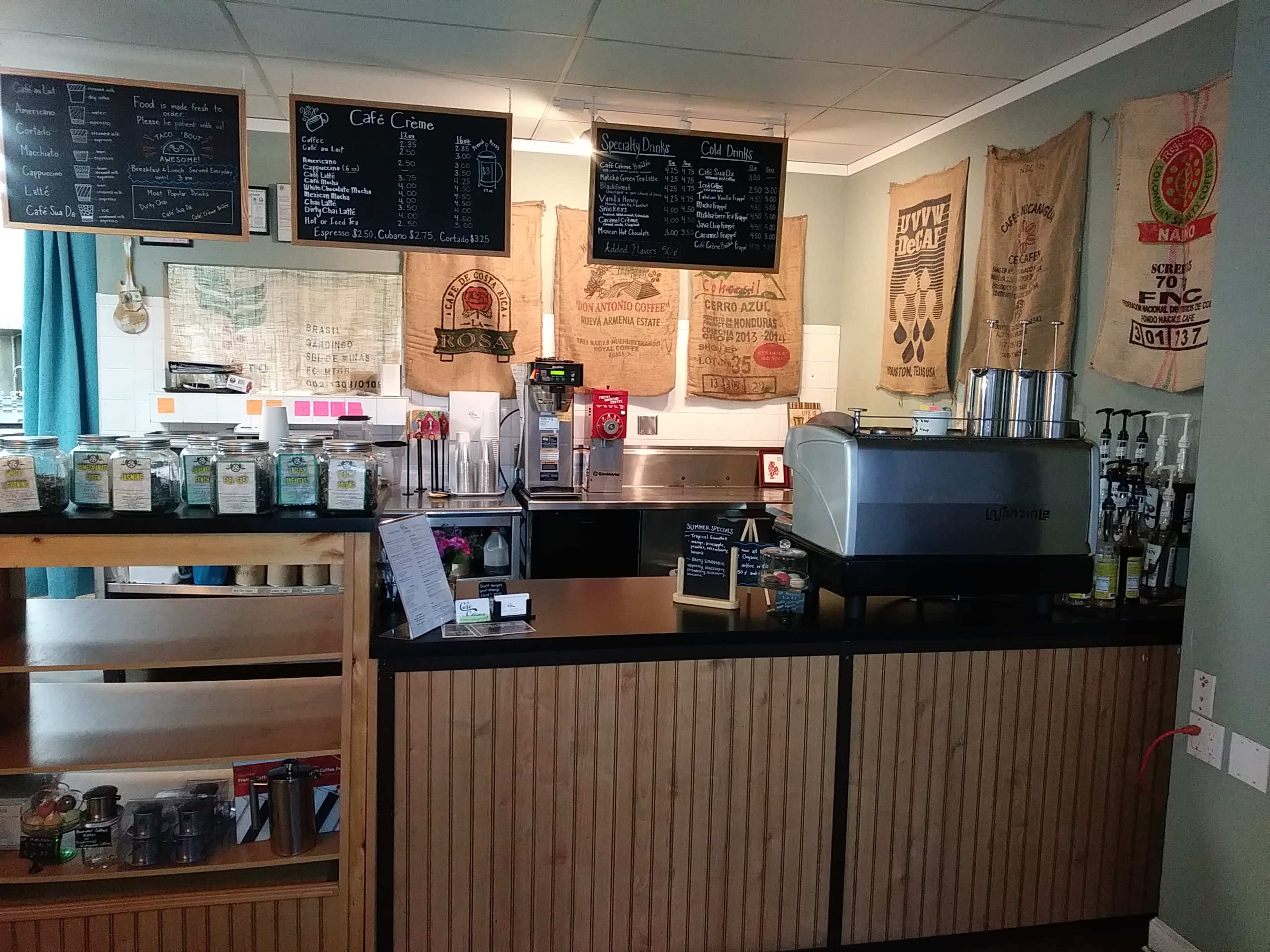 The image shows a coffee shop counter featuring a coffee machine, various drink menu items on chalkboards, and bags of coffee bean sacks hanging on the walls.