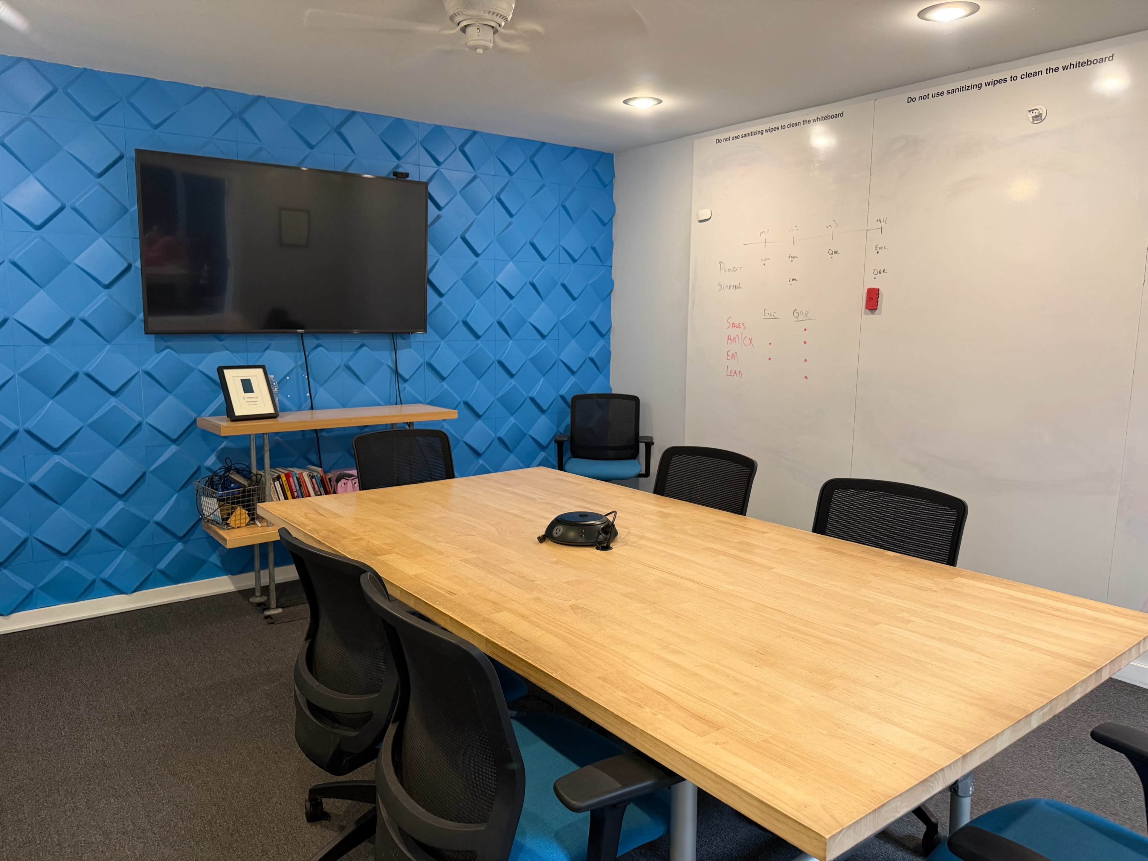 The image shows a modern conference room with a long wooden table, black office chairs, a wall-mounted TV, and a whiteboard on a blue textured wall.