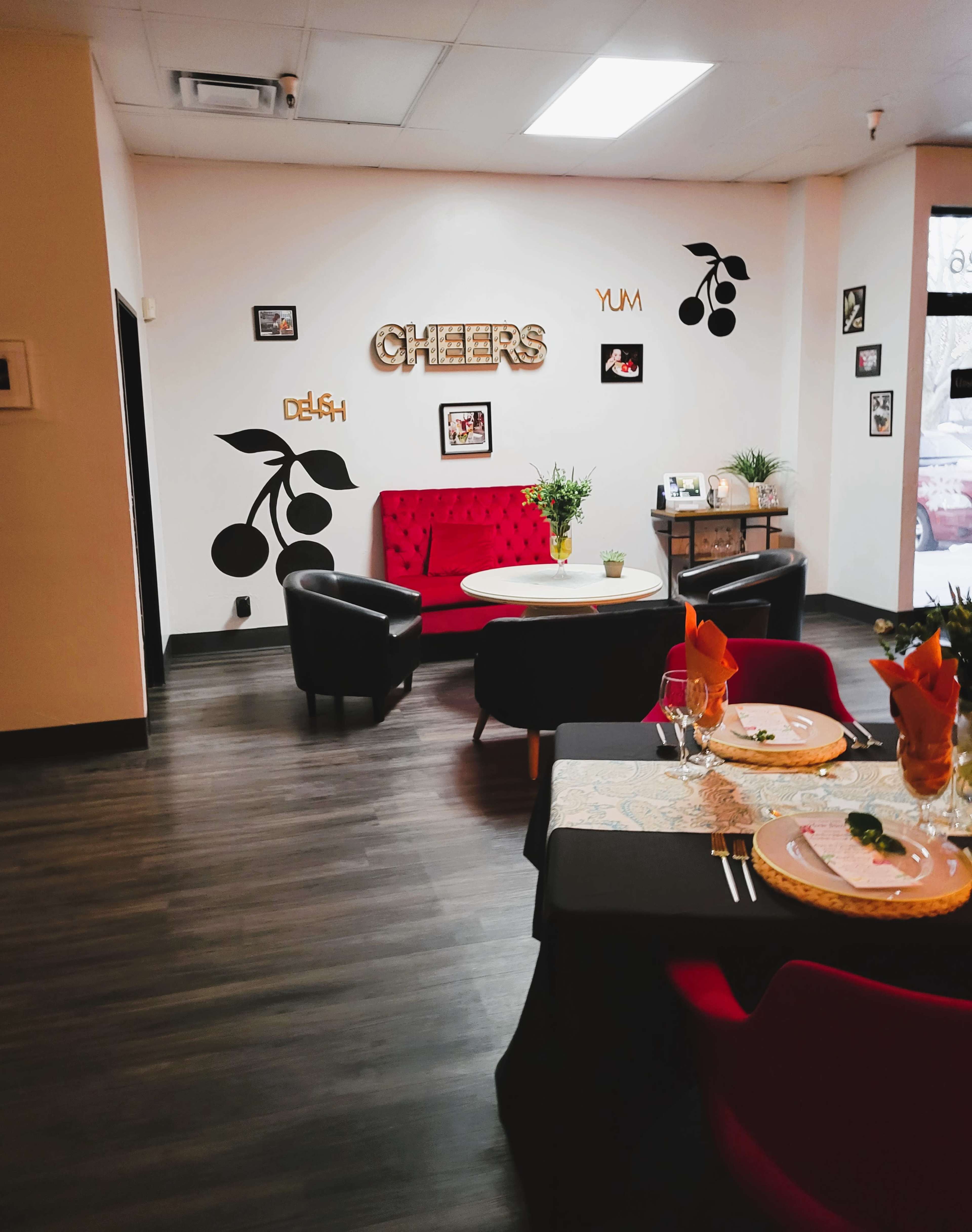 A cozy dining area features a red velvet sofa, elegant tables set with flowers, and wall decorations that include the words "CHEERS" and cherry motifs.
