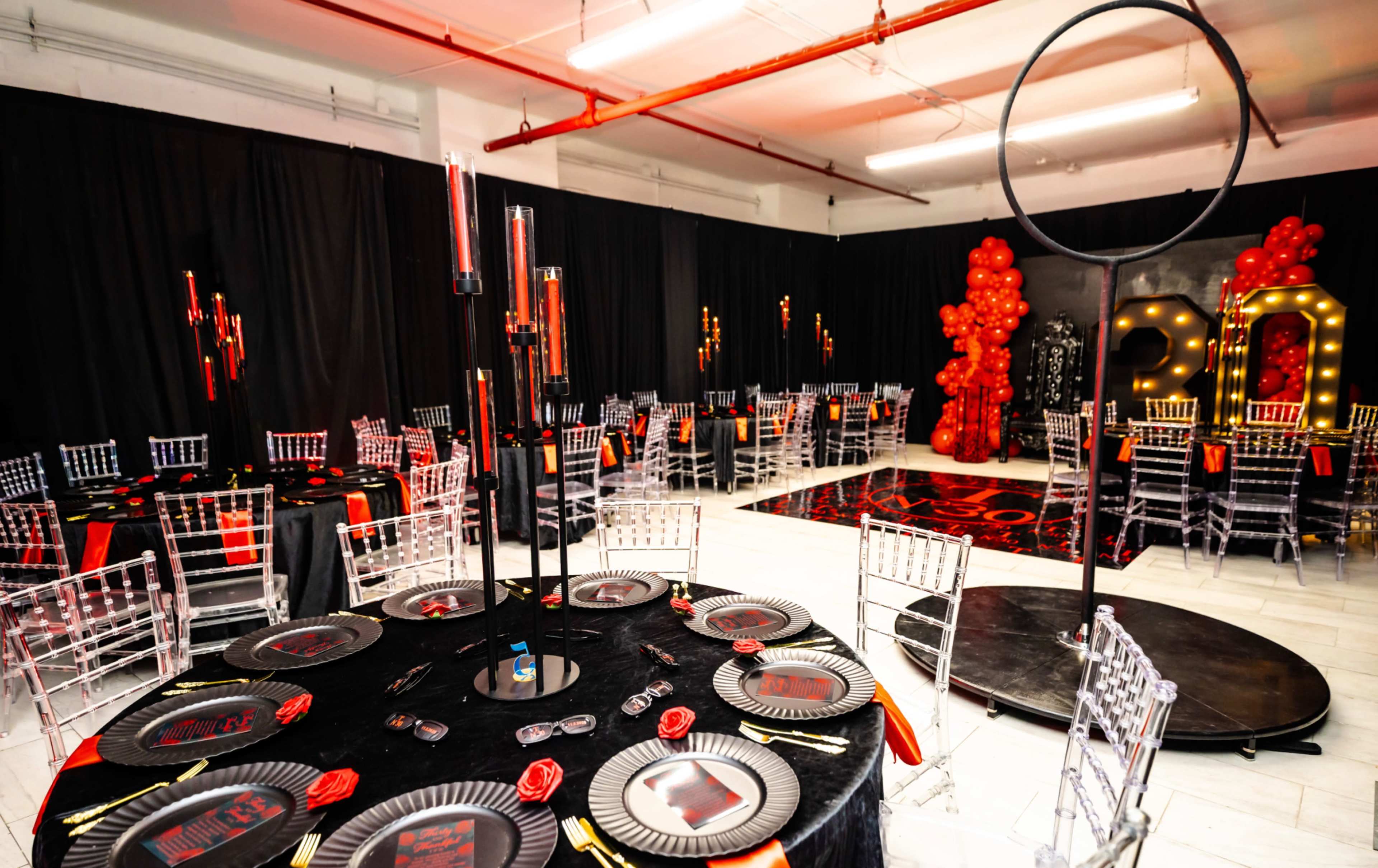 The image shows a decorated event space featuring black and red themed tables with elegant centerpieces, surrounded by chairs and a large backdrop illuminated with lights.