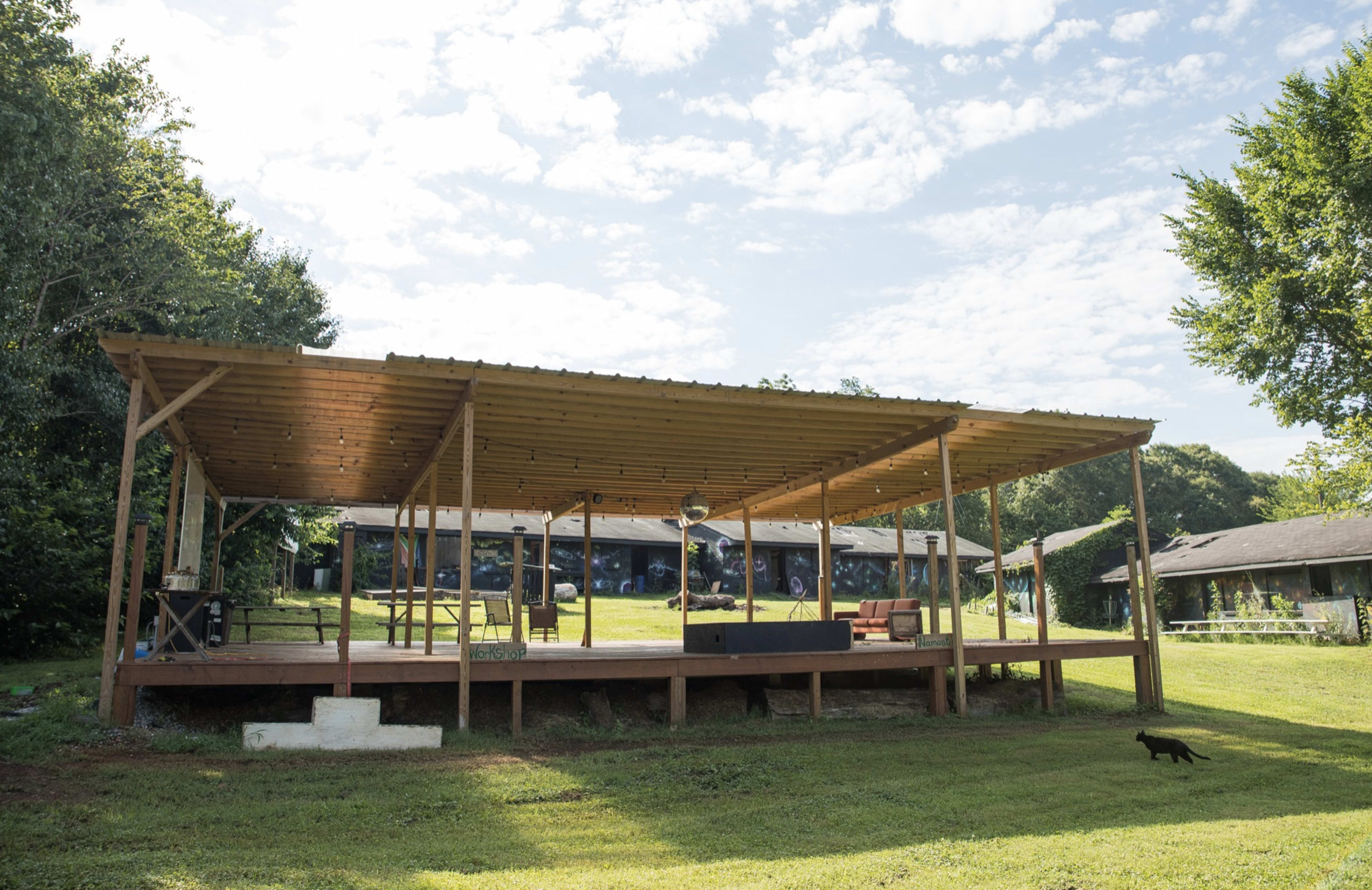 A wooden pavilion with a slatted roof stands on a grassy area, with benches and a small stage in front, and a black cat walking nearby.
