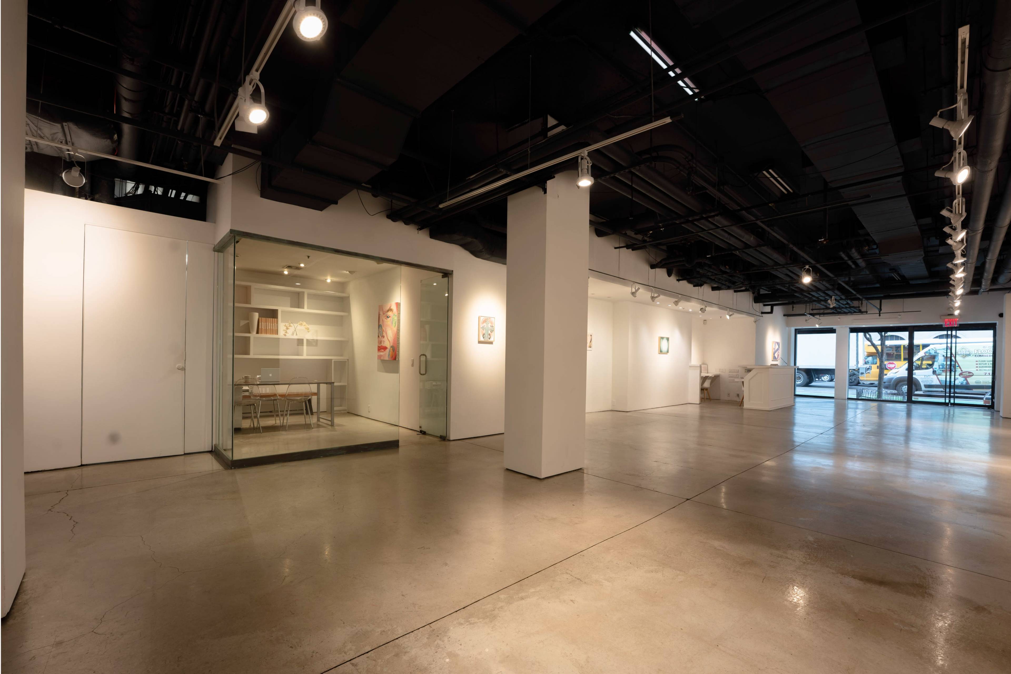 The image shows a spacious, modern art gallery featuring white walls, polished concrete floors, and a glass-enclosed room with a table and chairs.