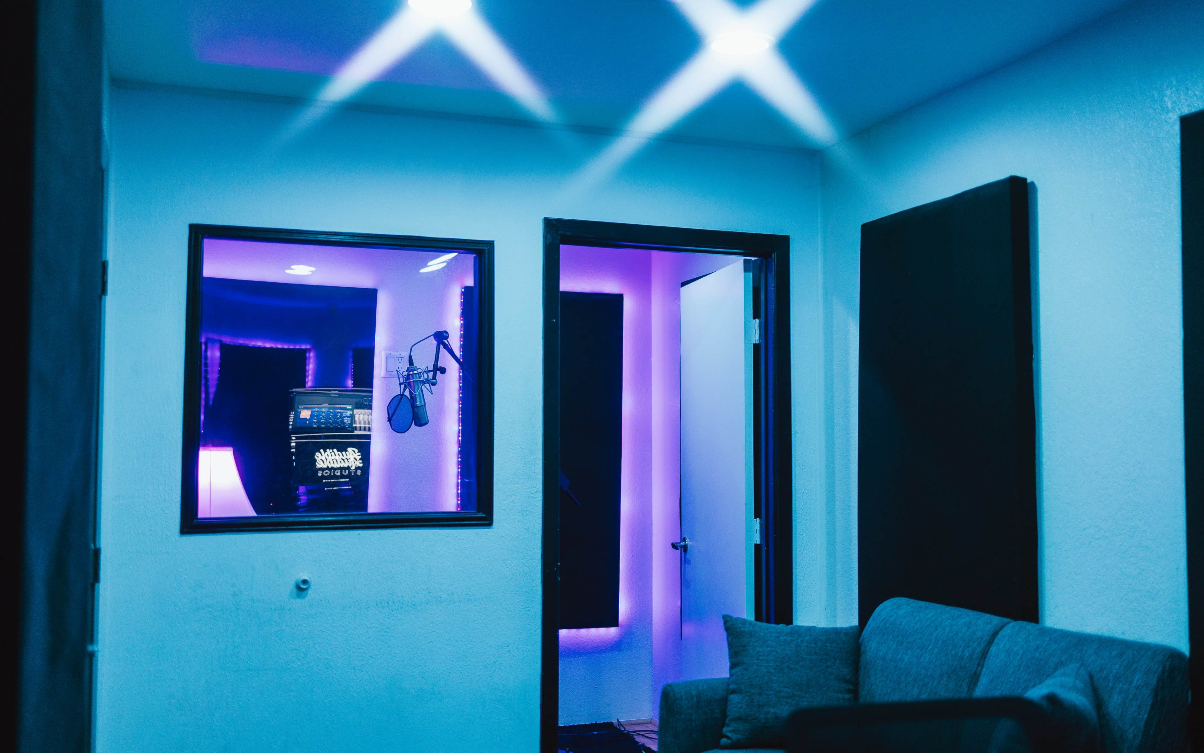 A music recording studio features a microphone setup visible through a glass window, with purple lighting and a cozy seating area.