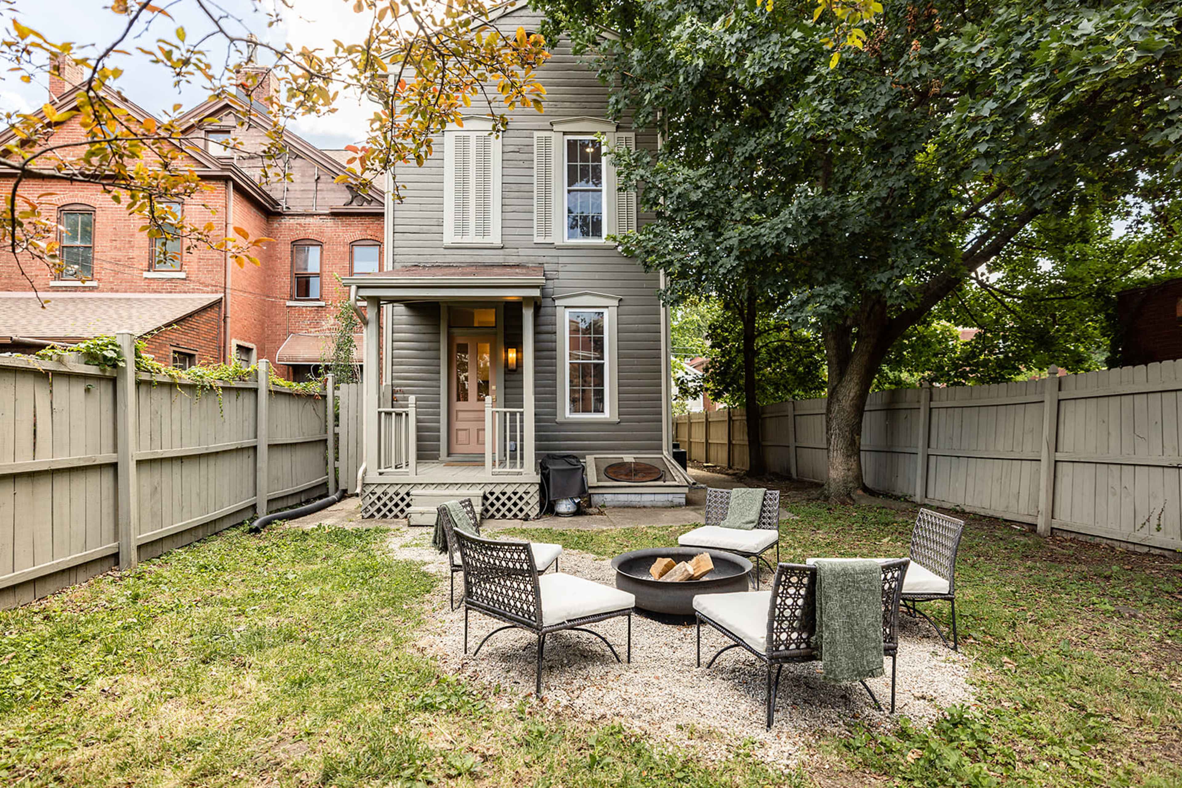 A three-story gray house with a fenced backyard features a fire pit surrounded by six chairs on a gravel patio.