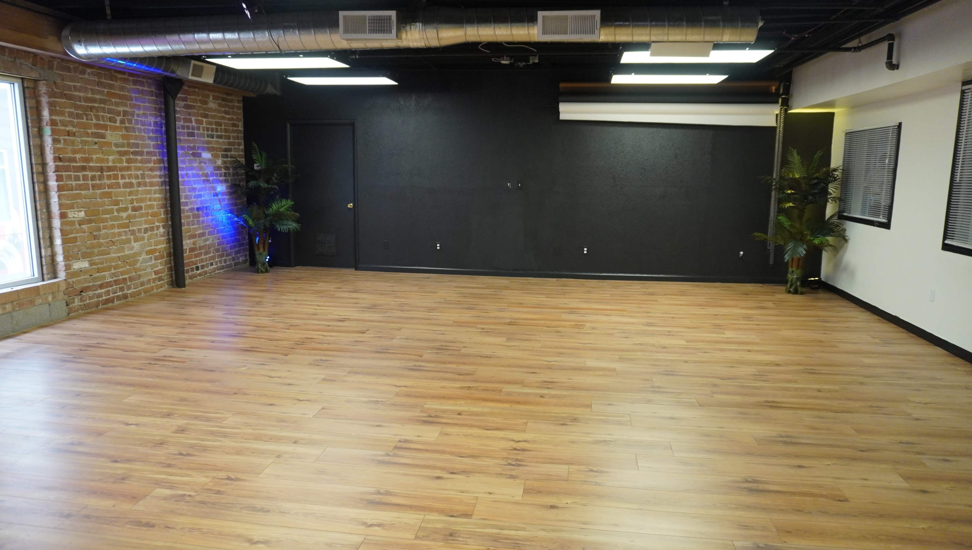 An empty studio space with wooden flooring, exposed brick walls, and a dark accent wall, supplemented by a few potted plants near the corners.