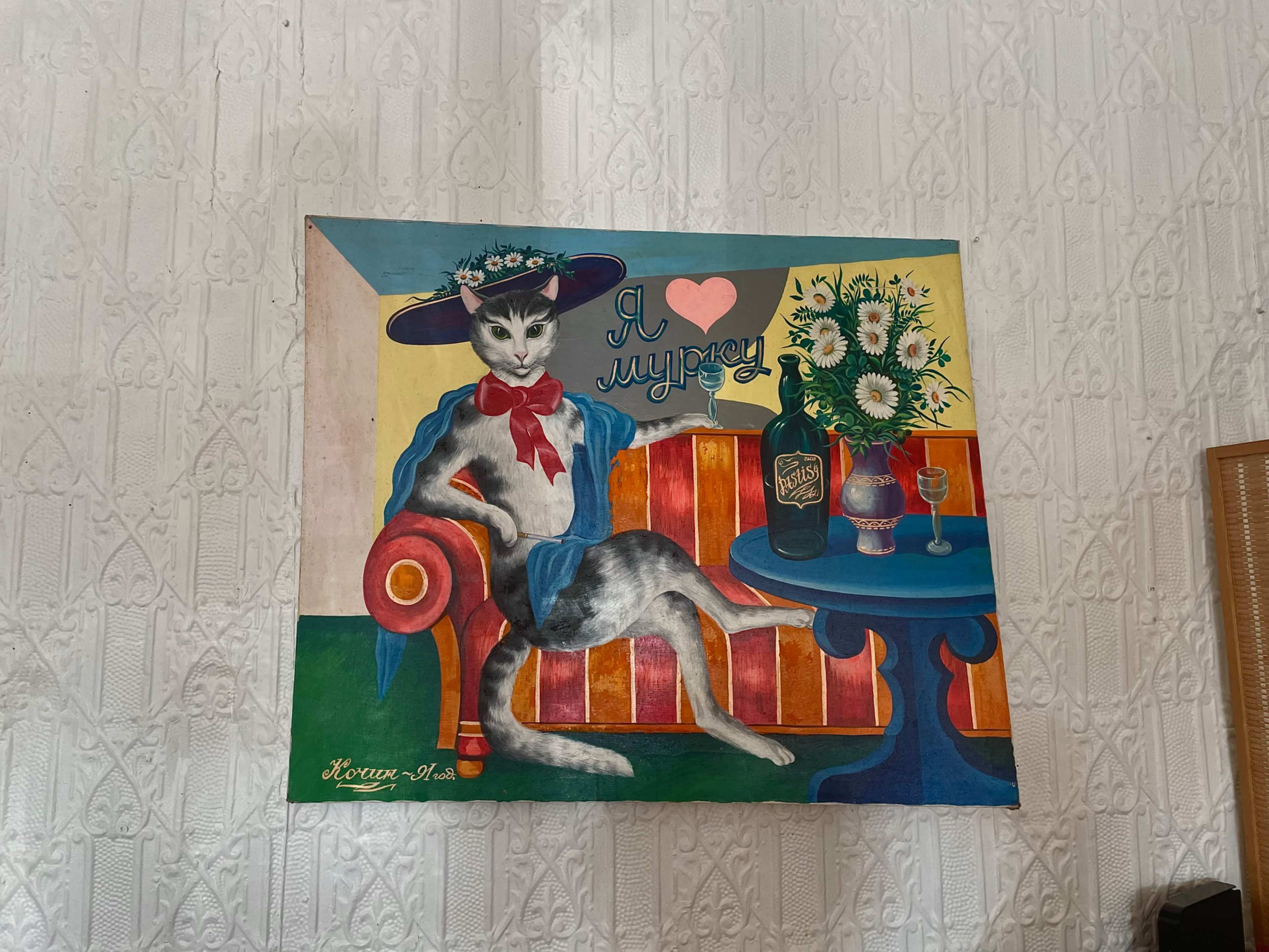 The image features a colorful painting of a cat dressed in a blue outfit and a wide-brimmed hat, sitting on a red and blue striped couch beside a table with flowers and drinks.