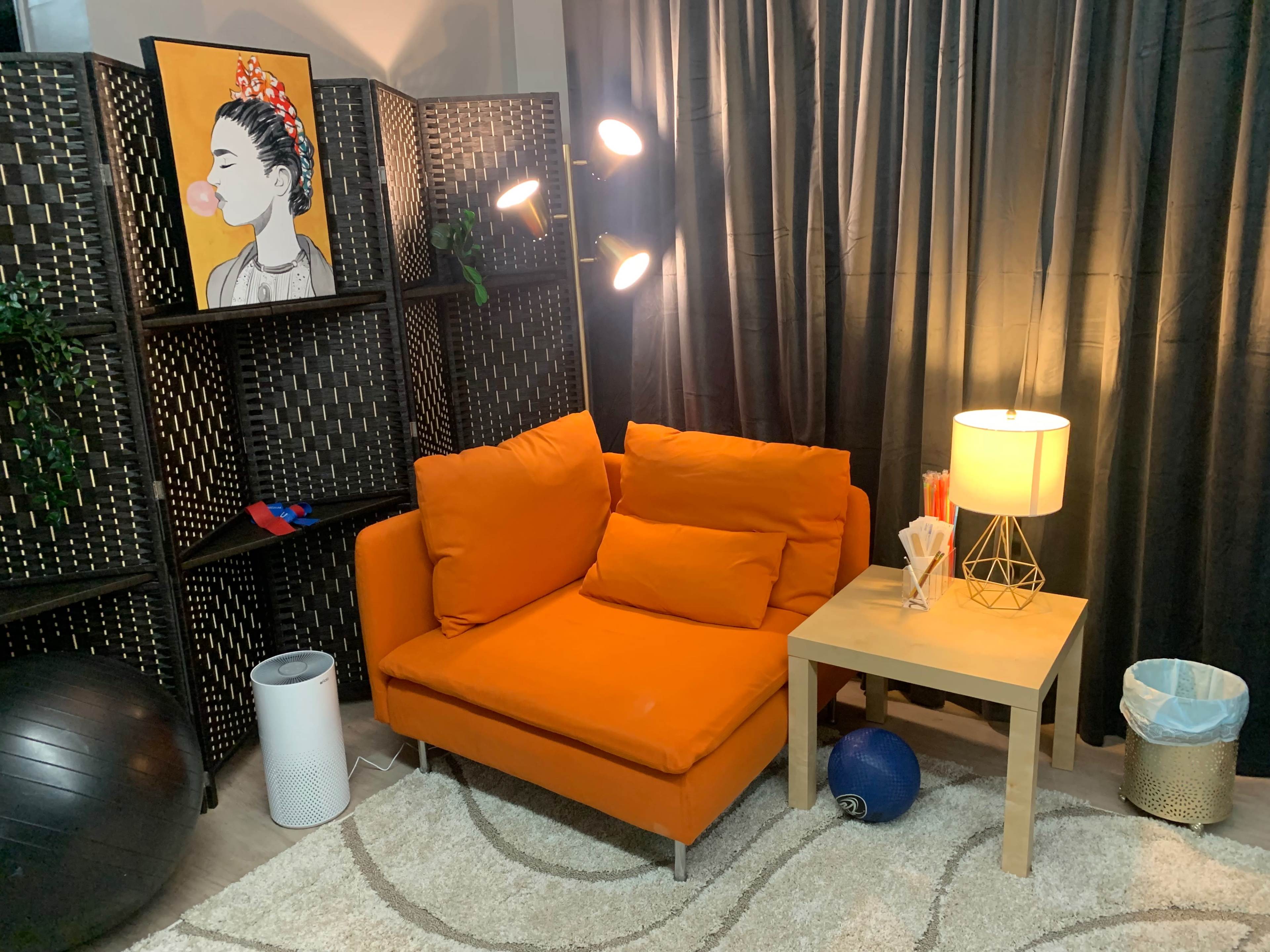 A small living area with an orange couch, a side table with a lamp, and an artwork on the wall, all surrounded by black curtains and a woven screen.