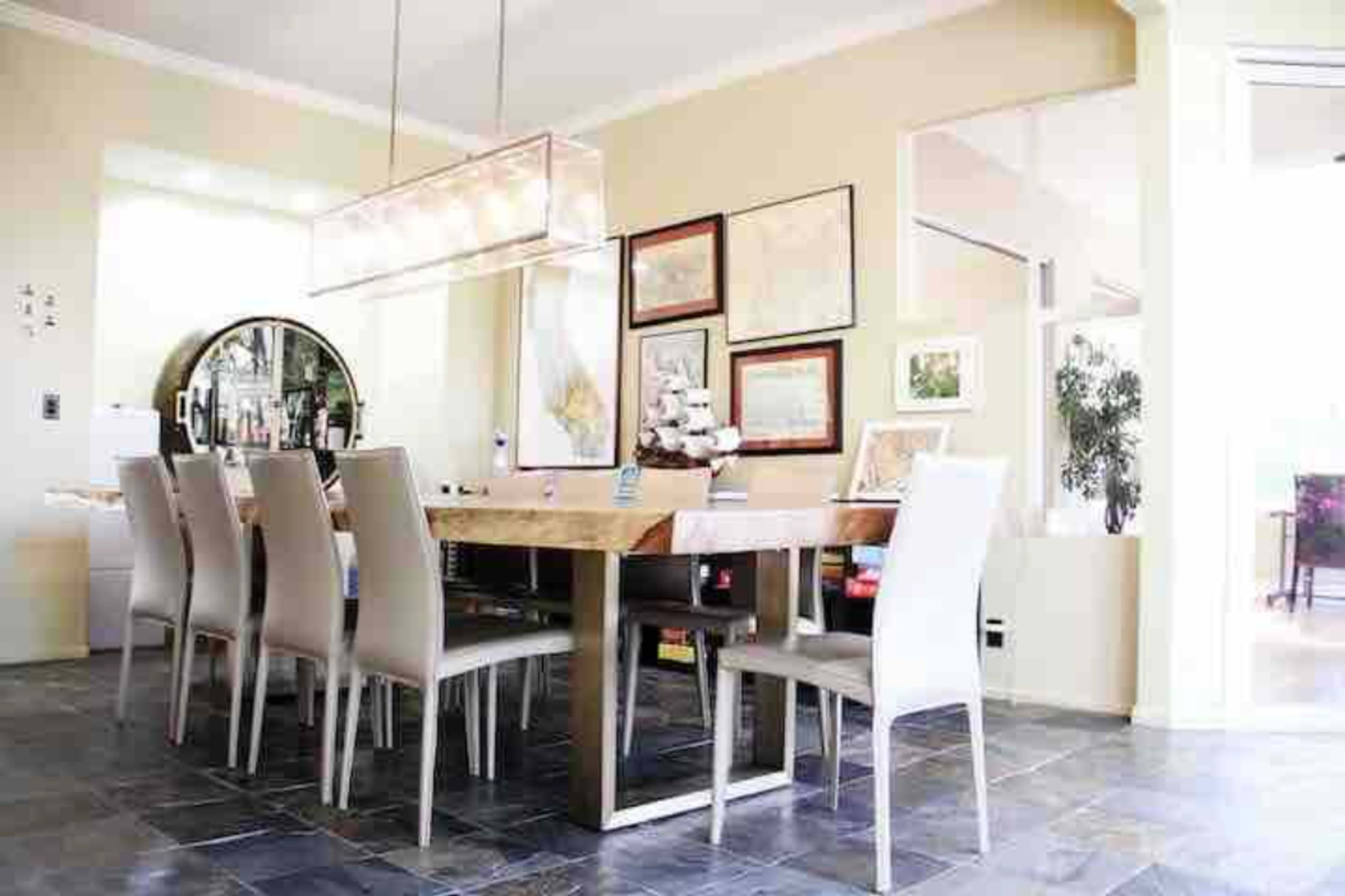 A spacious dining area features a long wooden table surrounded by eight light-colored chairs, with a large pendant light overhead and framed artwork on the walls.