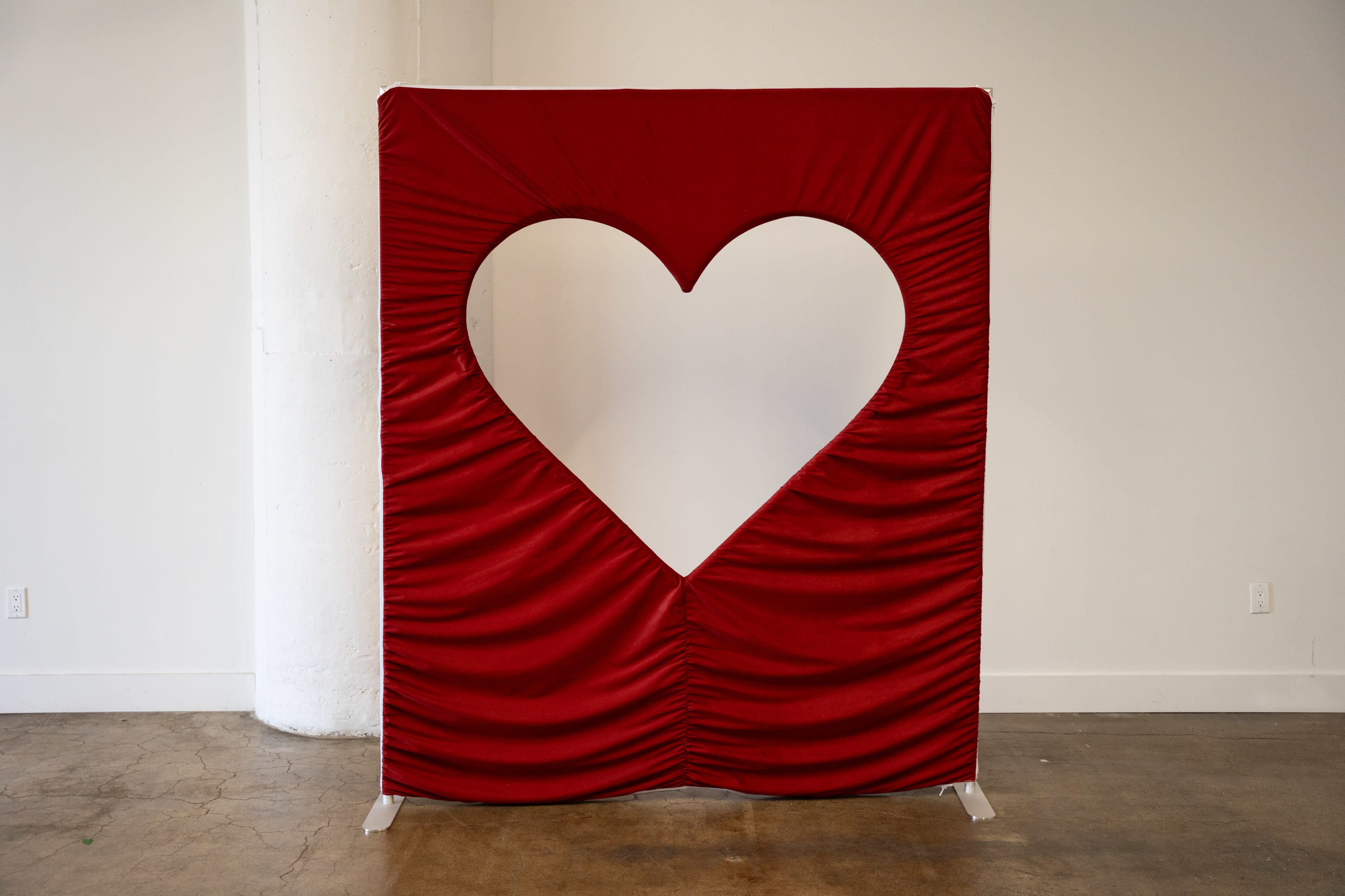 The image shows a large, red fabric display with a heart-shaped cutout, standing against a white wall in a minimalist space.