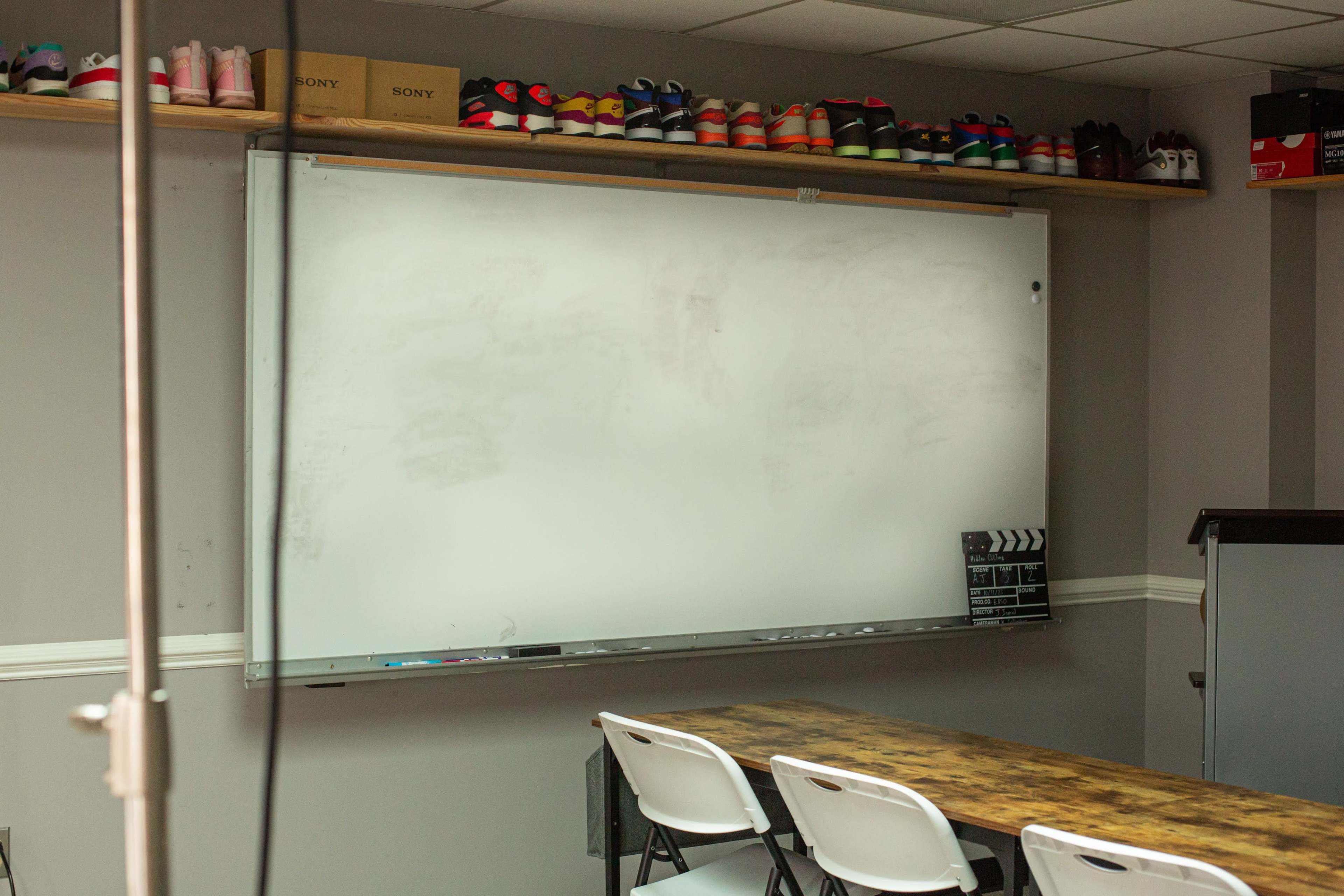 A classroom with a large whiteboard mounted on the wall and a wooden table with white chairs in front of it, while various shoes are neatly arranged on a shelf above.