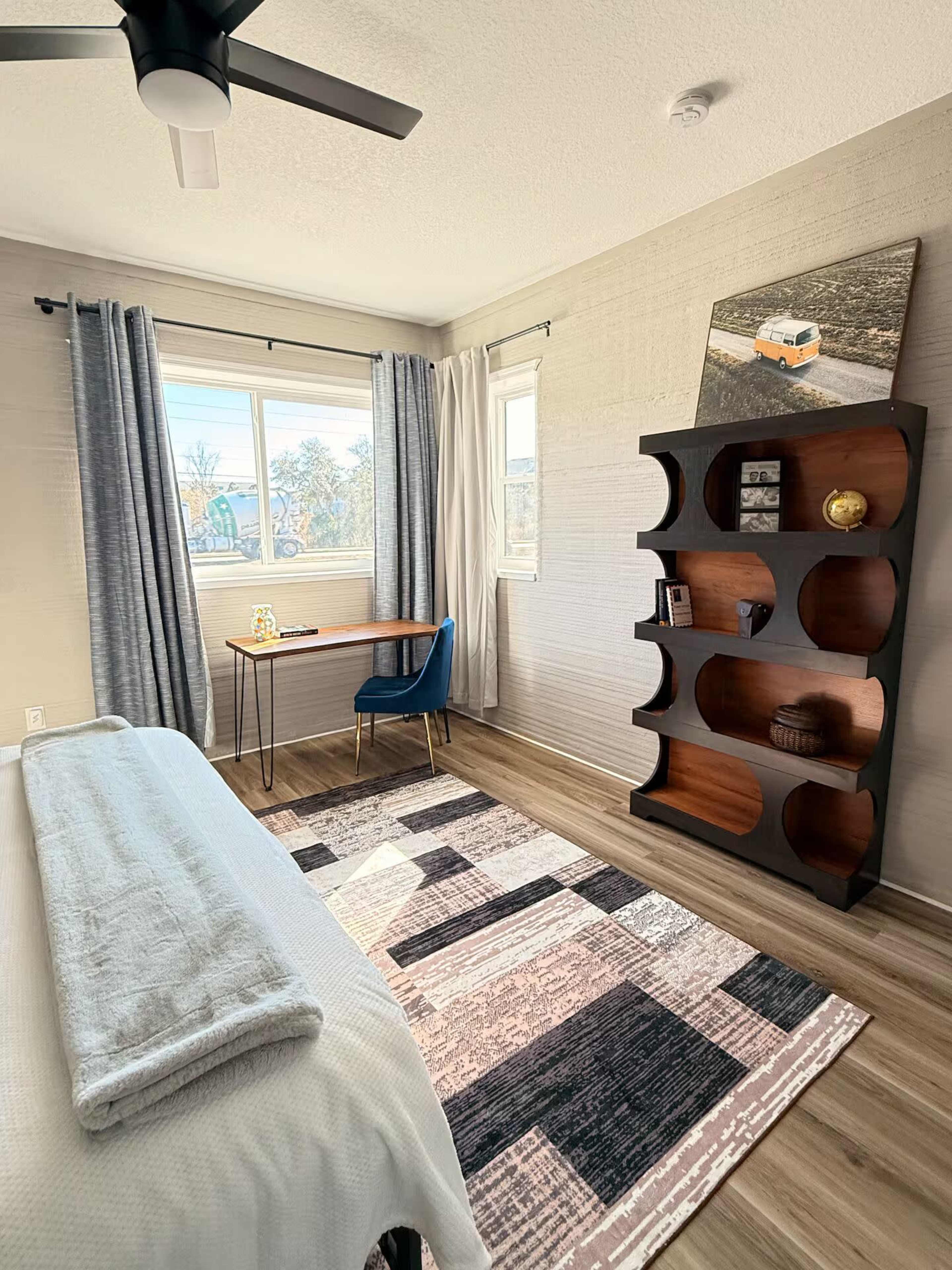 The image shows a neatly arranged bedroom featuring a bed, a small desk with a chair, a bookshelf, large windows with curtains, and a patterned rug on the floor.