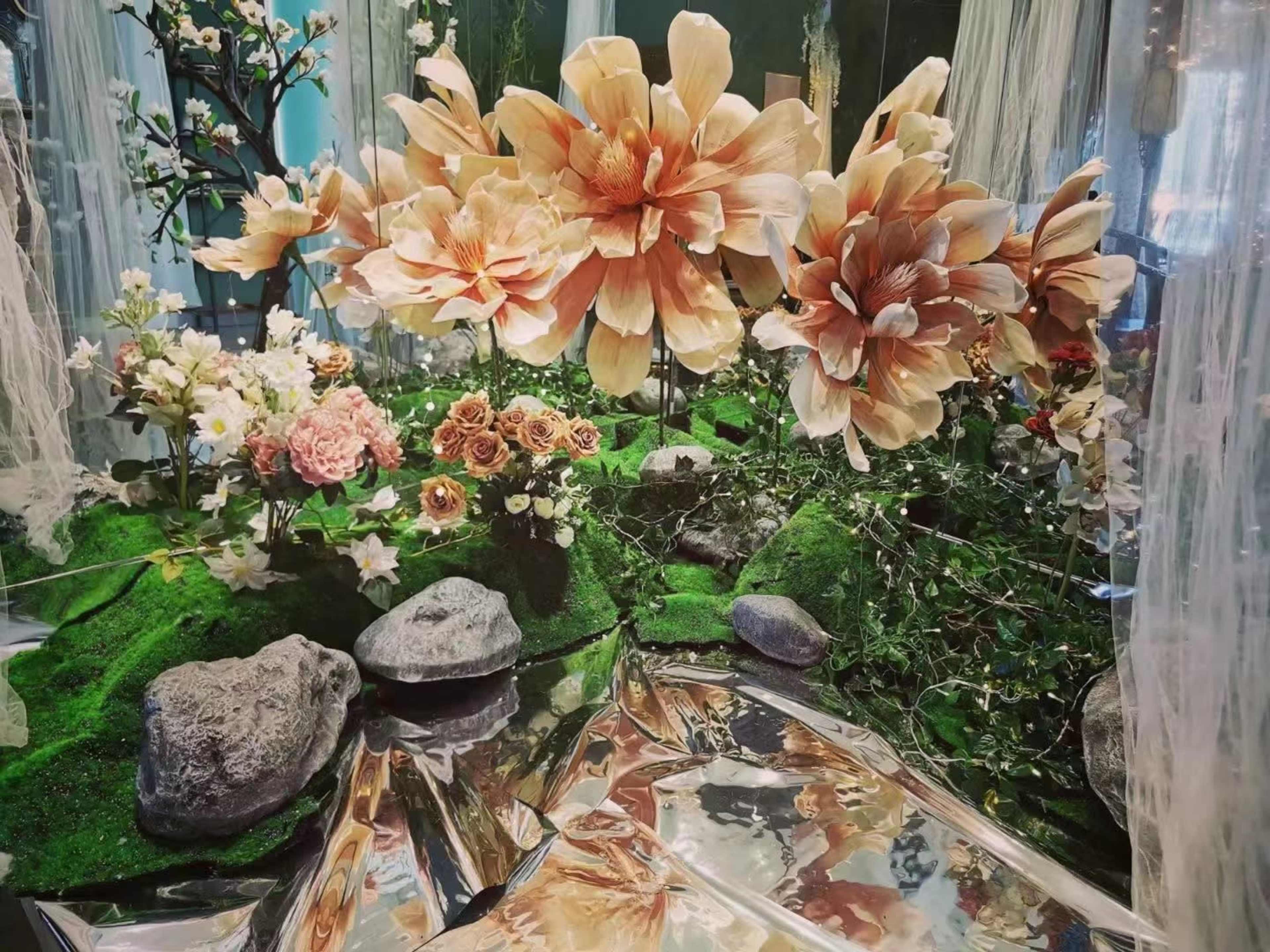 A display of large artificial flowers and smaller plants arranged among stones on a reflective surface.