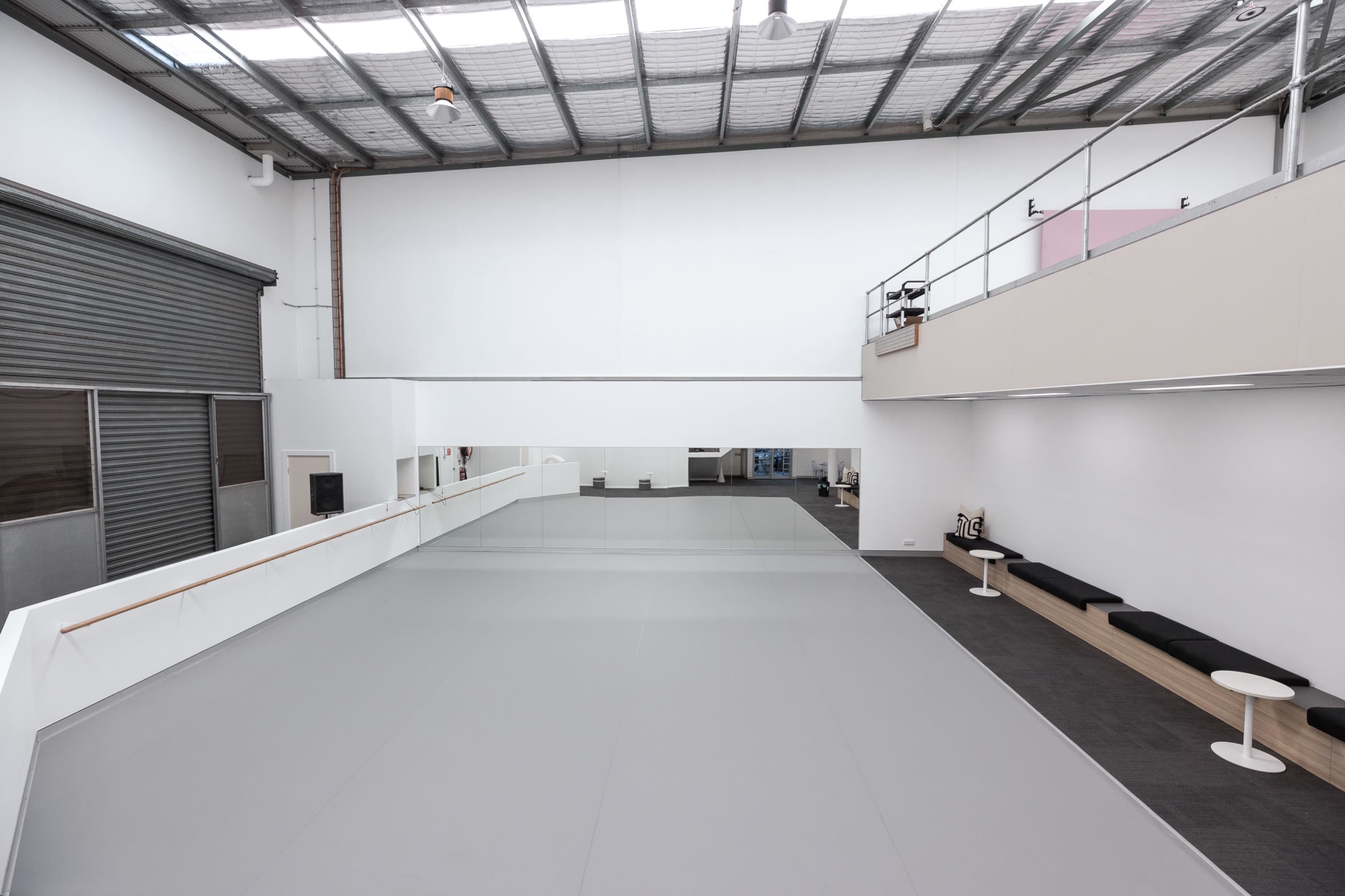 A spacious, well-lit dance studio featuring a large gray dance floor and a mirrored wall, with seating along one side.