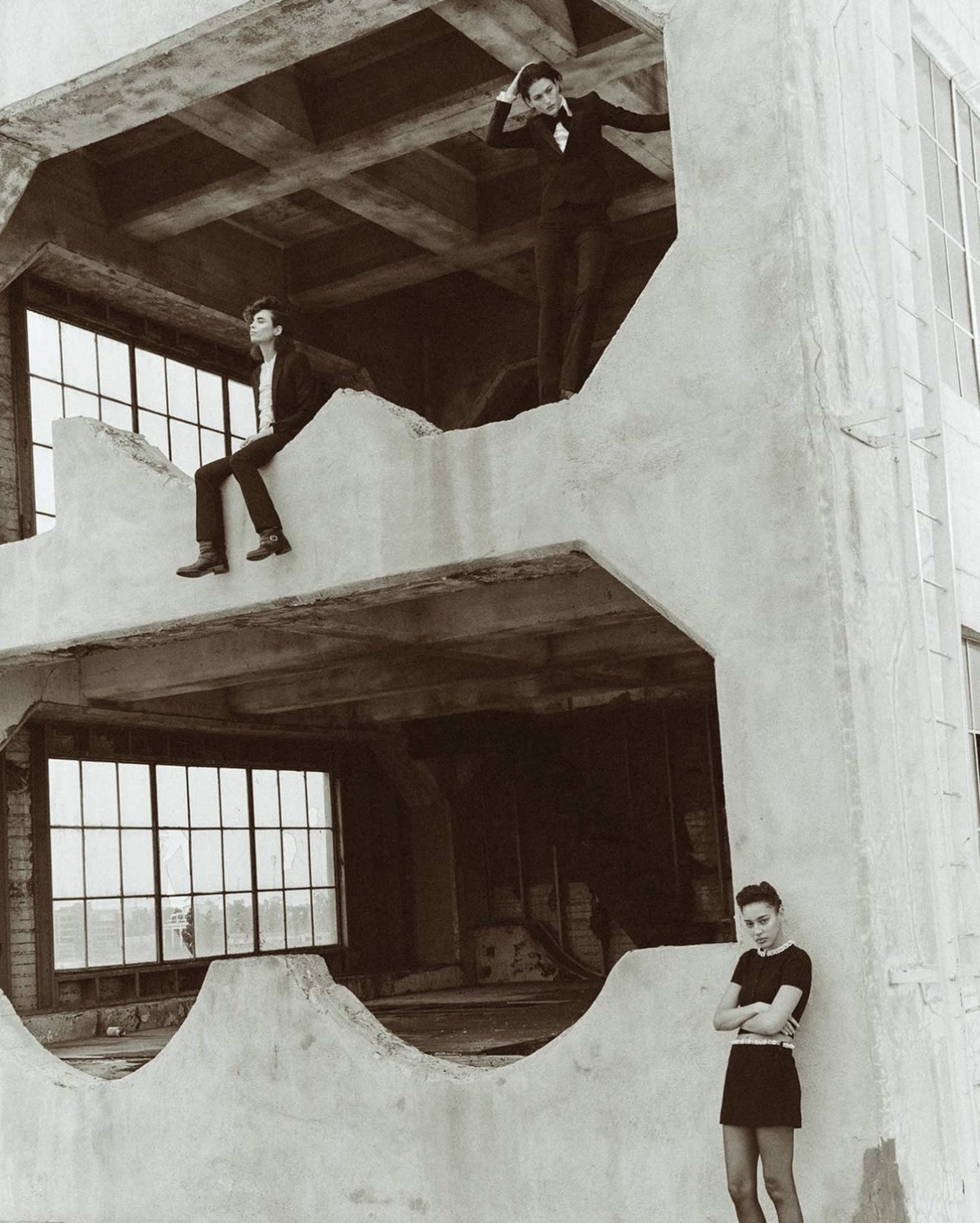 Three models in black outfits pose around a partially demolished building with large windows and exposed concrete.