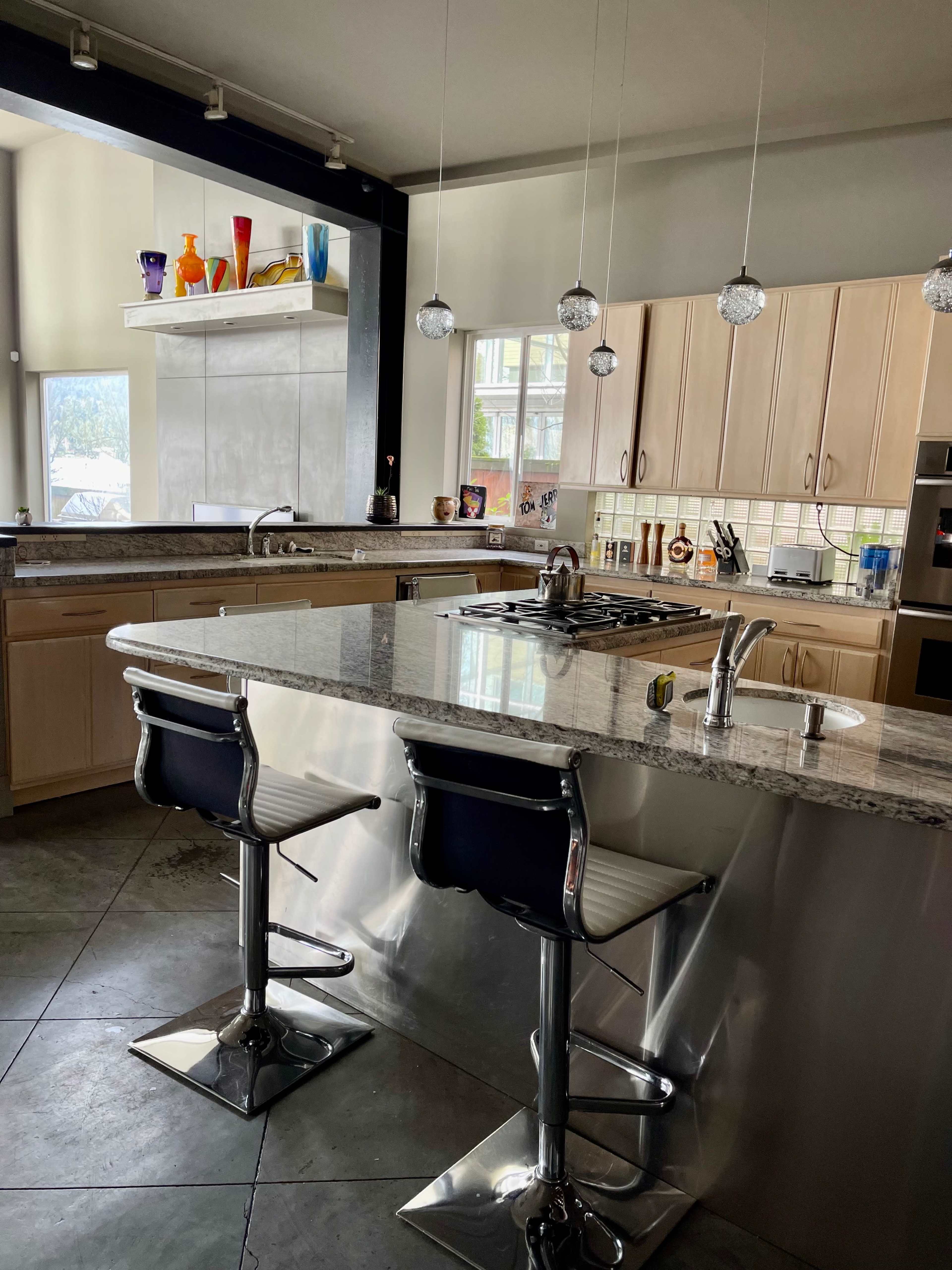 A modern kitchen with a granite island featuring two chrome bar stools, stainless steel appliances, and large windows allowing natural light.