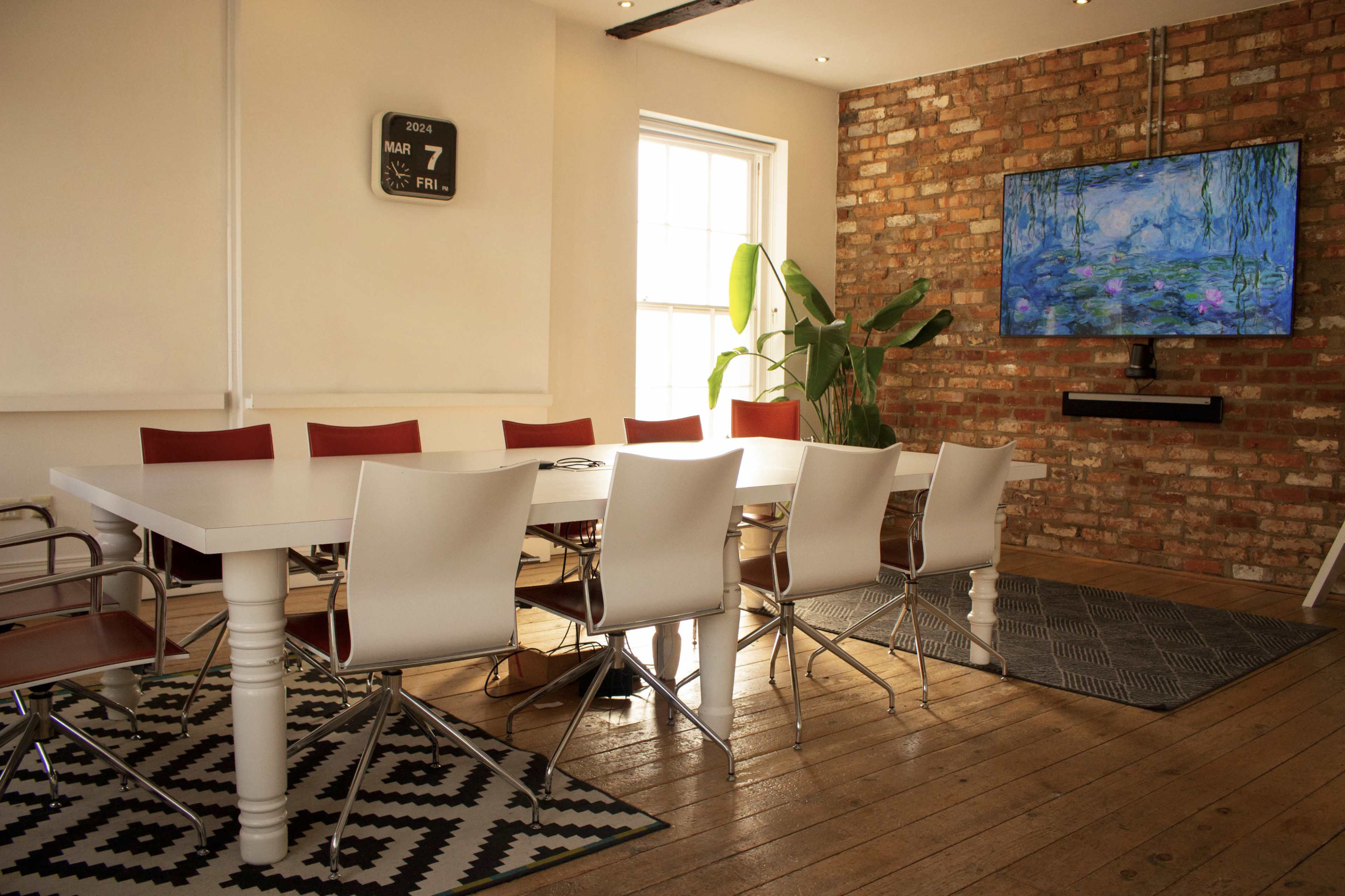 A modern meeting room features a white table surrounded by white and red chairs, with a large TV mounted on a brick wall and a plant in the corner.