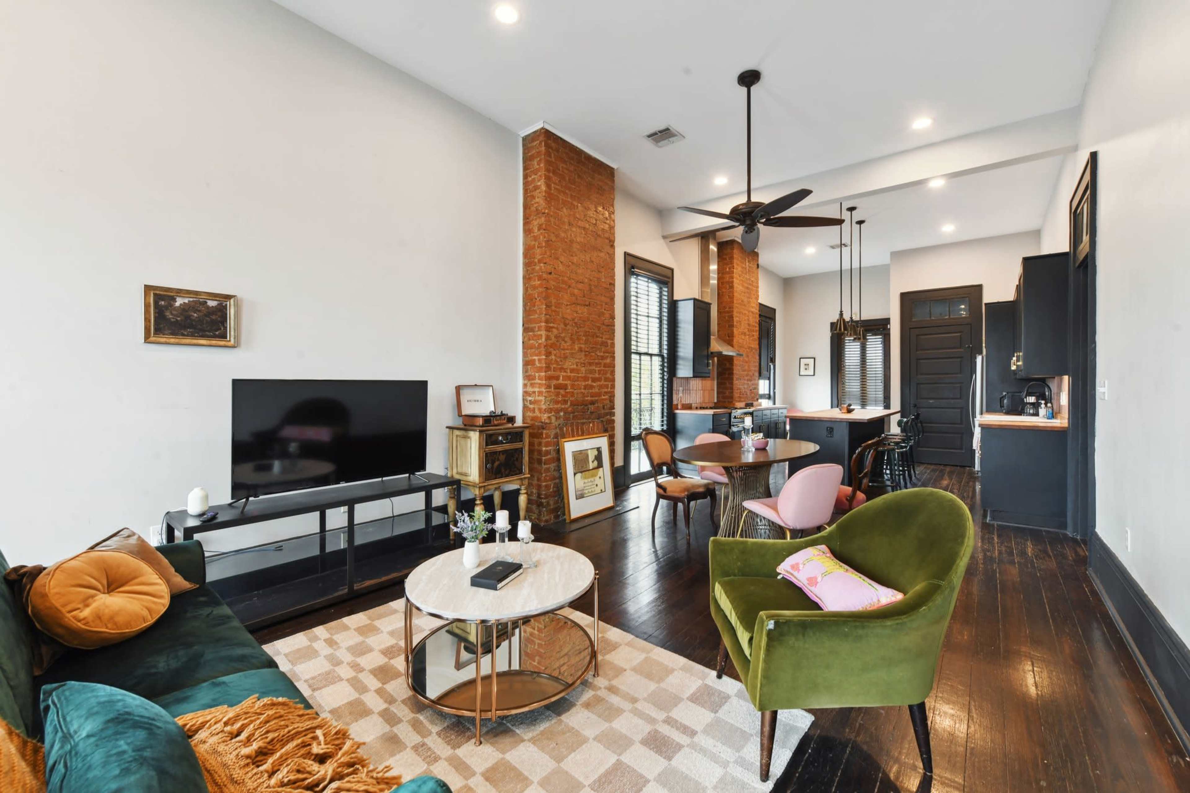A modern living room with a green sofa, a circular coffee table, a television on a stand, and a dining area featuring a kitchen in the background.