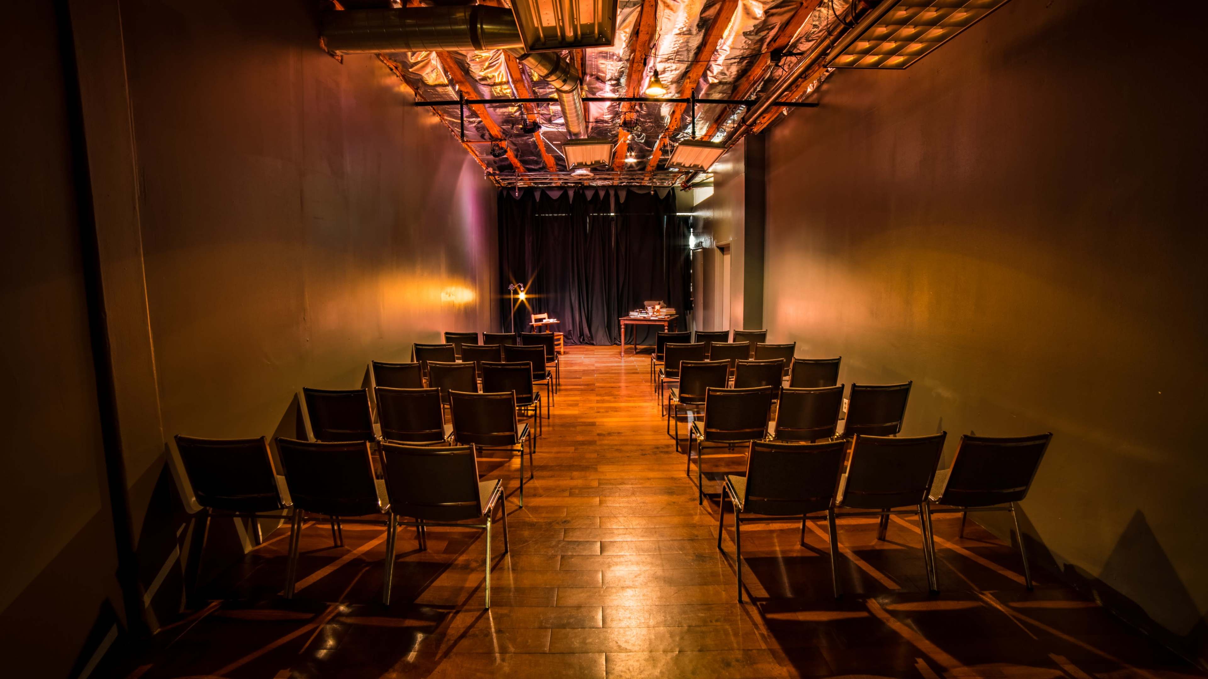 A dimly lit auditorium with rows of empty chairs facing a small stage at the end of a wooden floor.