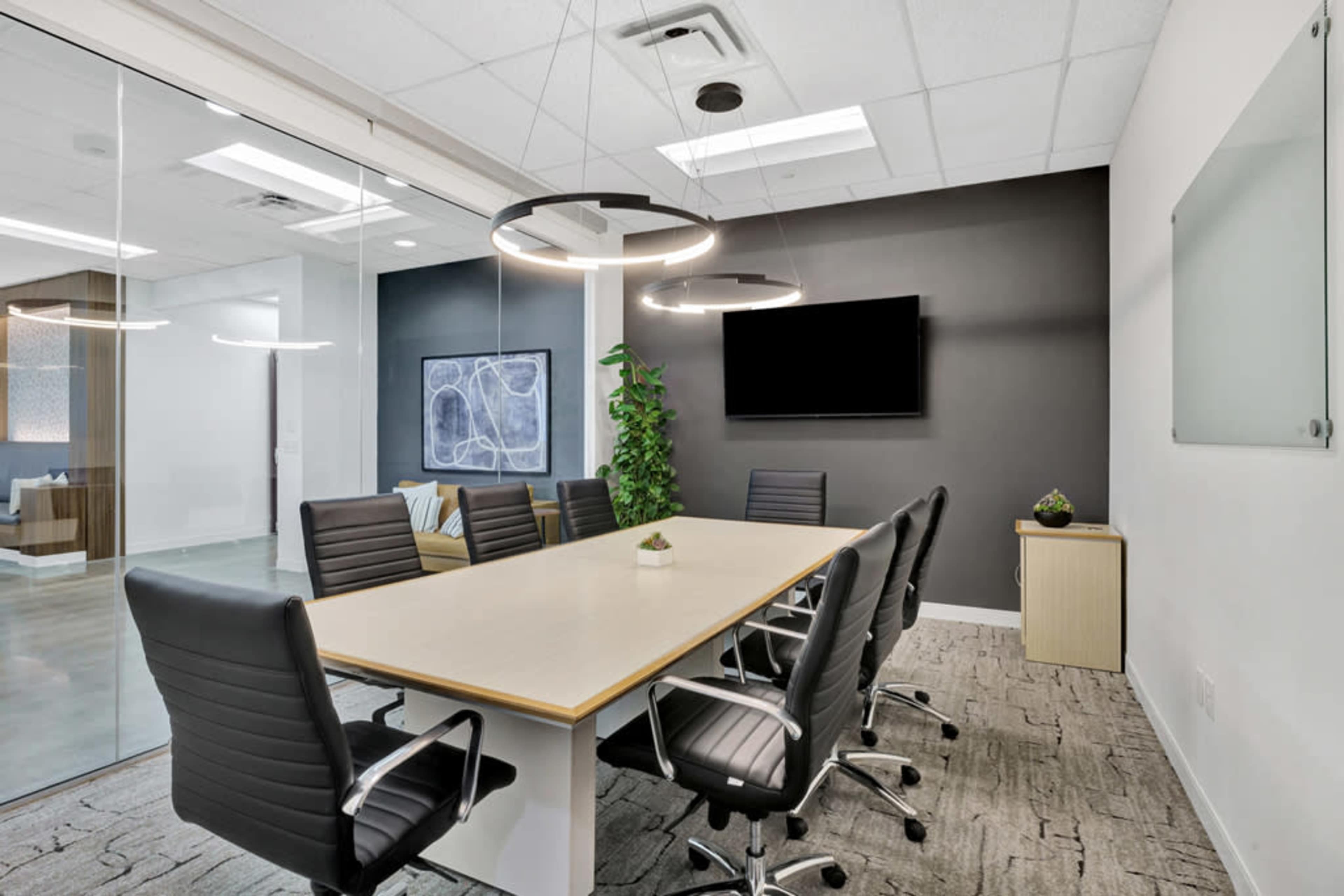 A modern conference room features a long table surrounded by black chairs, with a large screen on the wall and a potted plant in the corner.