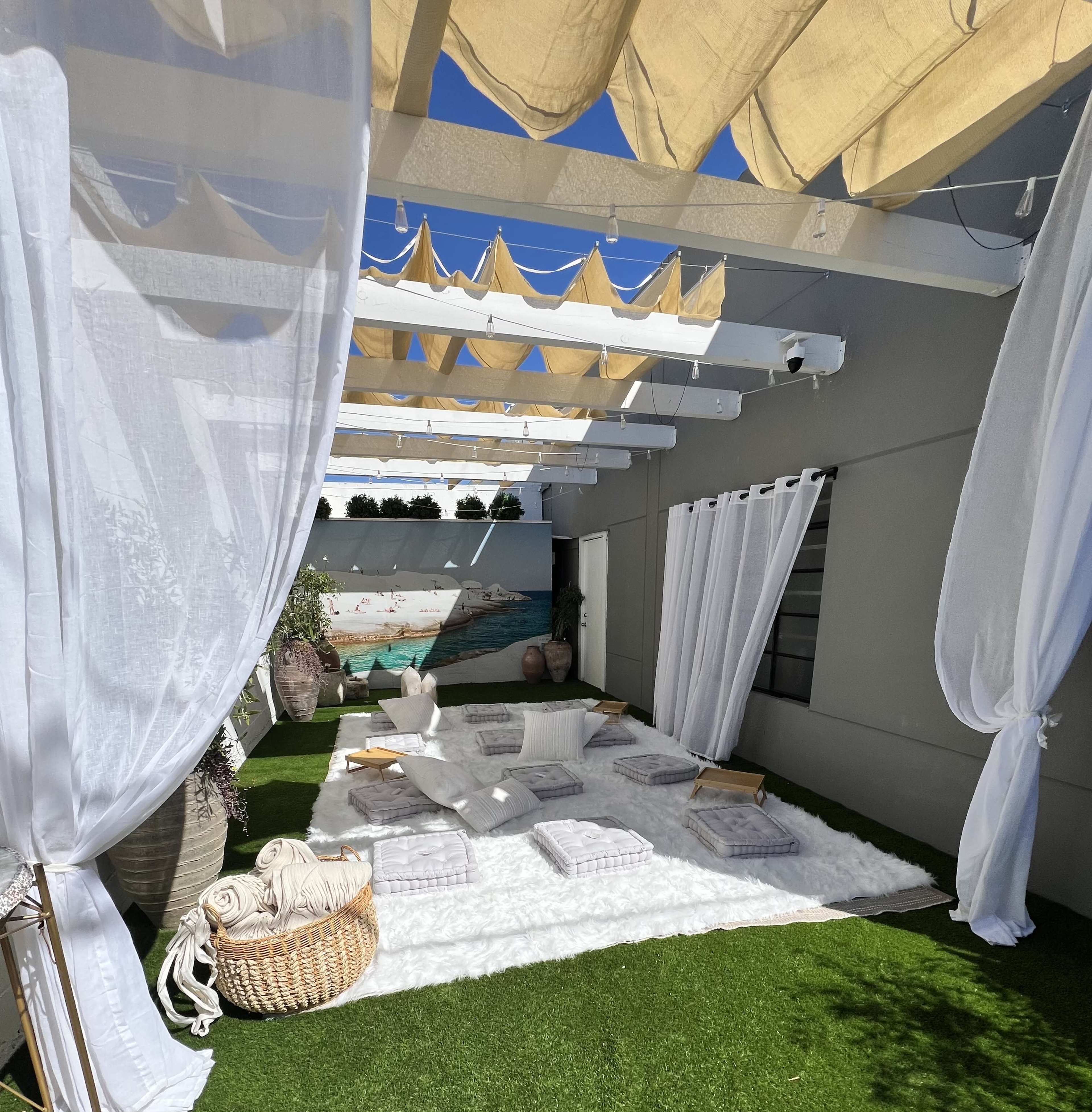 The image shows a serene outdoor space with white drapery, a soft white rug, wooden seating, and a mural depicting a beach scene.