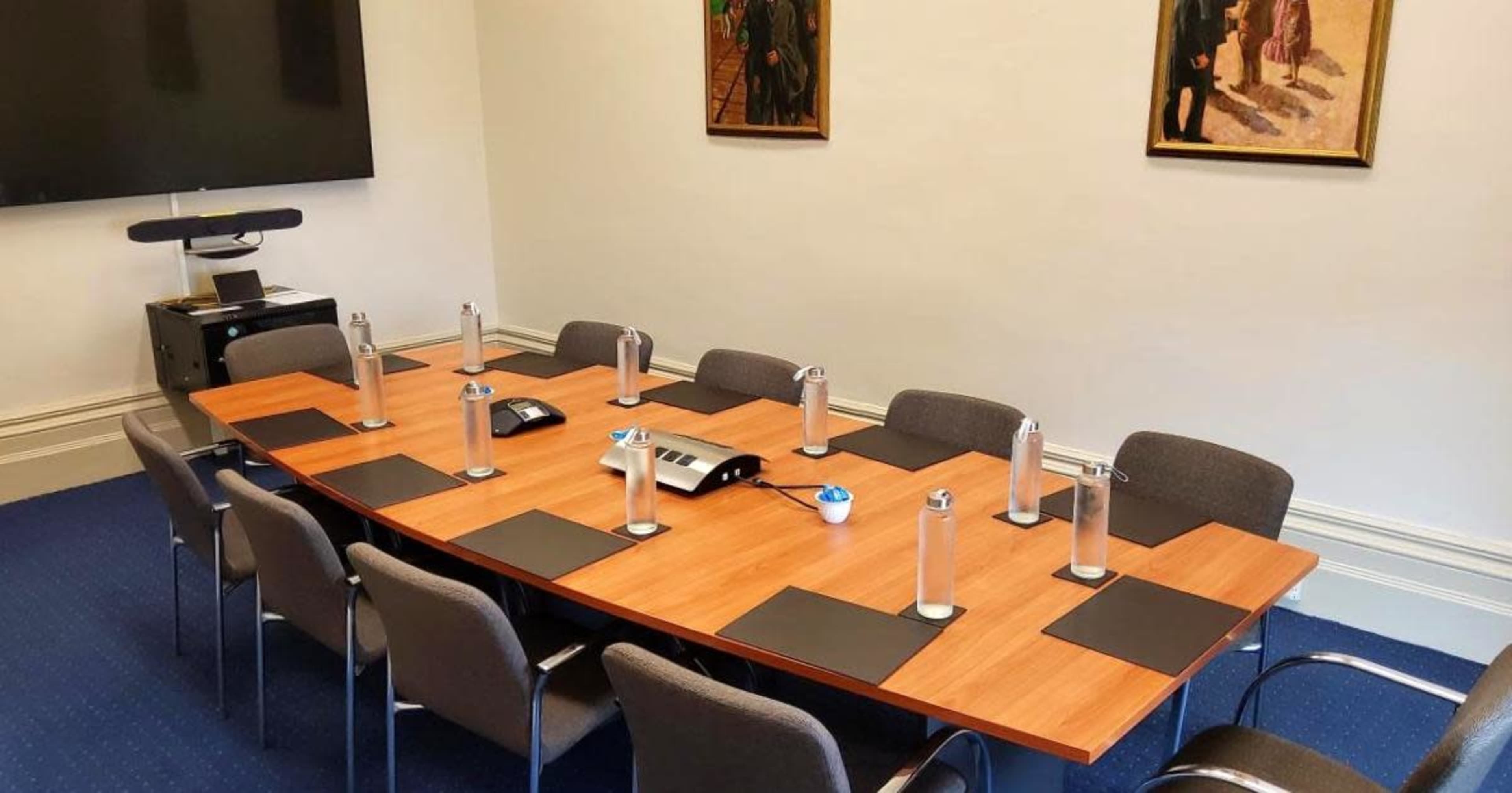 A large conference room features a wooden table surrounded by gray chairs, with water bottles and notepads positioned at each place setting, and two paintings hanging on the walls.