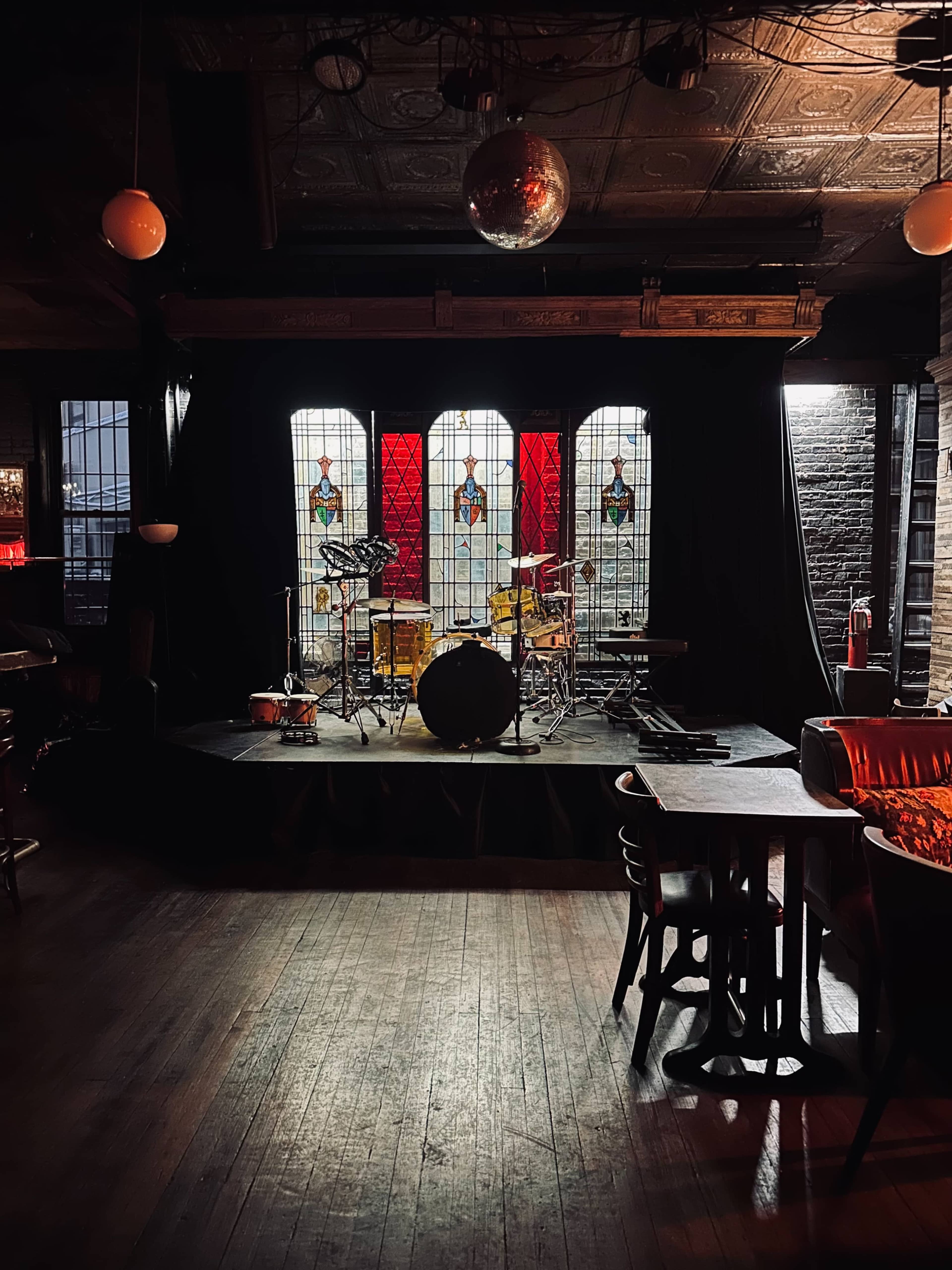 The image shows a dimly lit music stage in a venue, featuring a drum set positioned in front of stained glass windows.